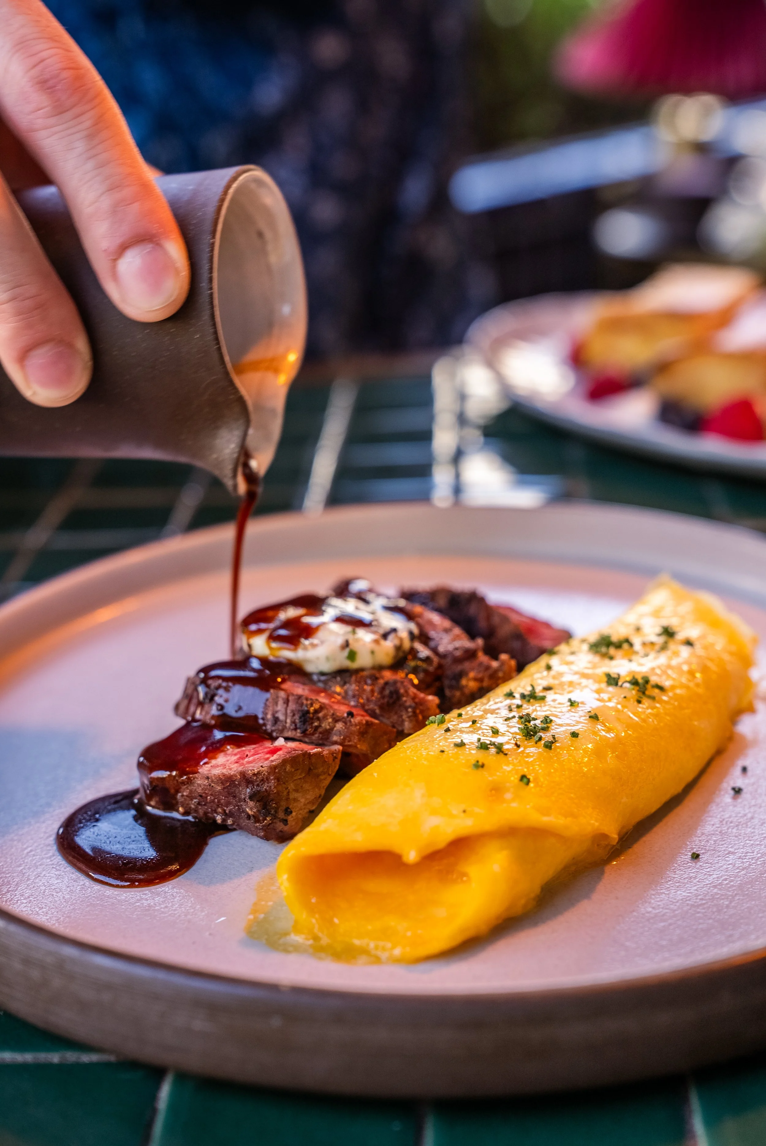 A plate of sliced steak topped with a dollop of butter and drizzled with sauce, served alongside a yellow omelette garnished with herbs.