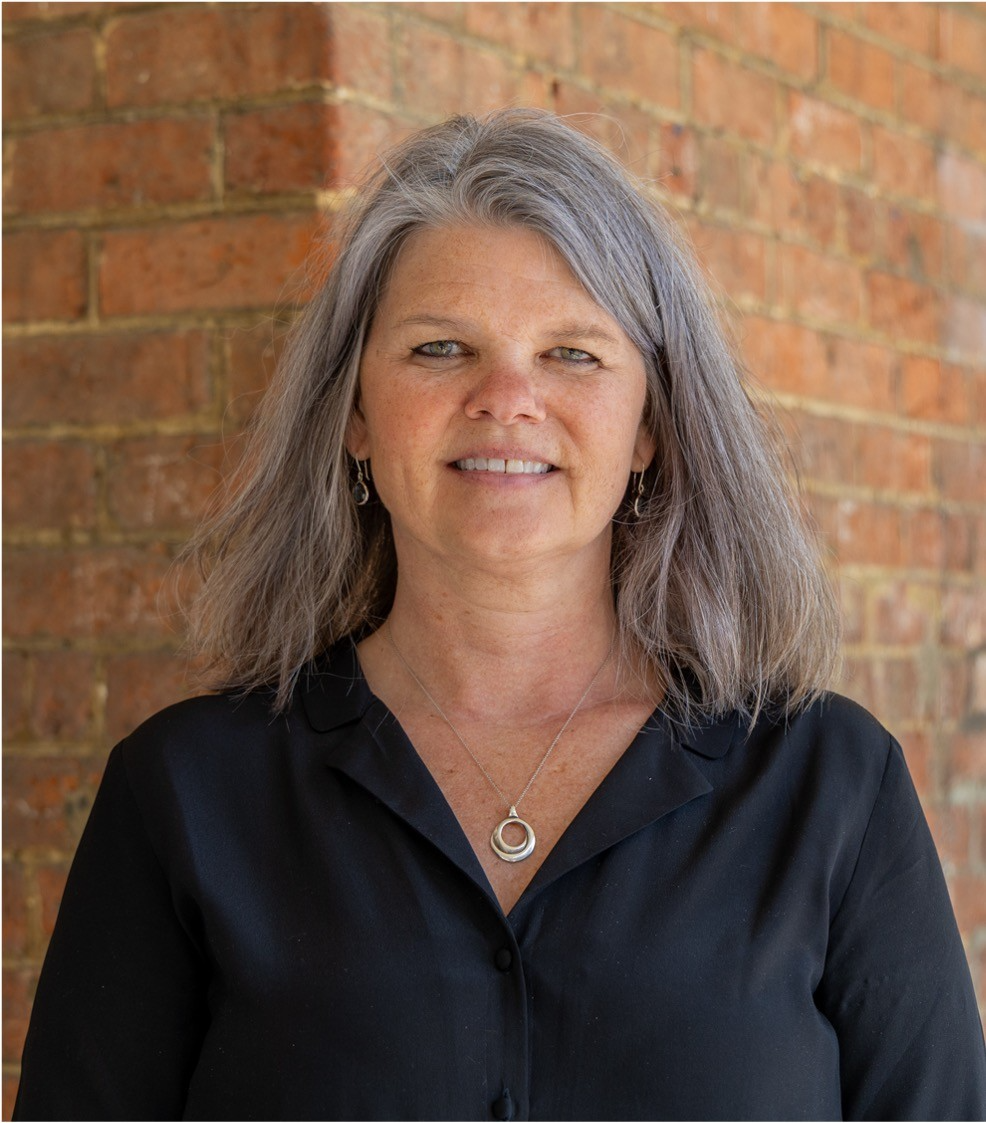 A woman with gray hair wearing a dark shirt, in front of a brick building background