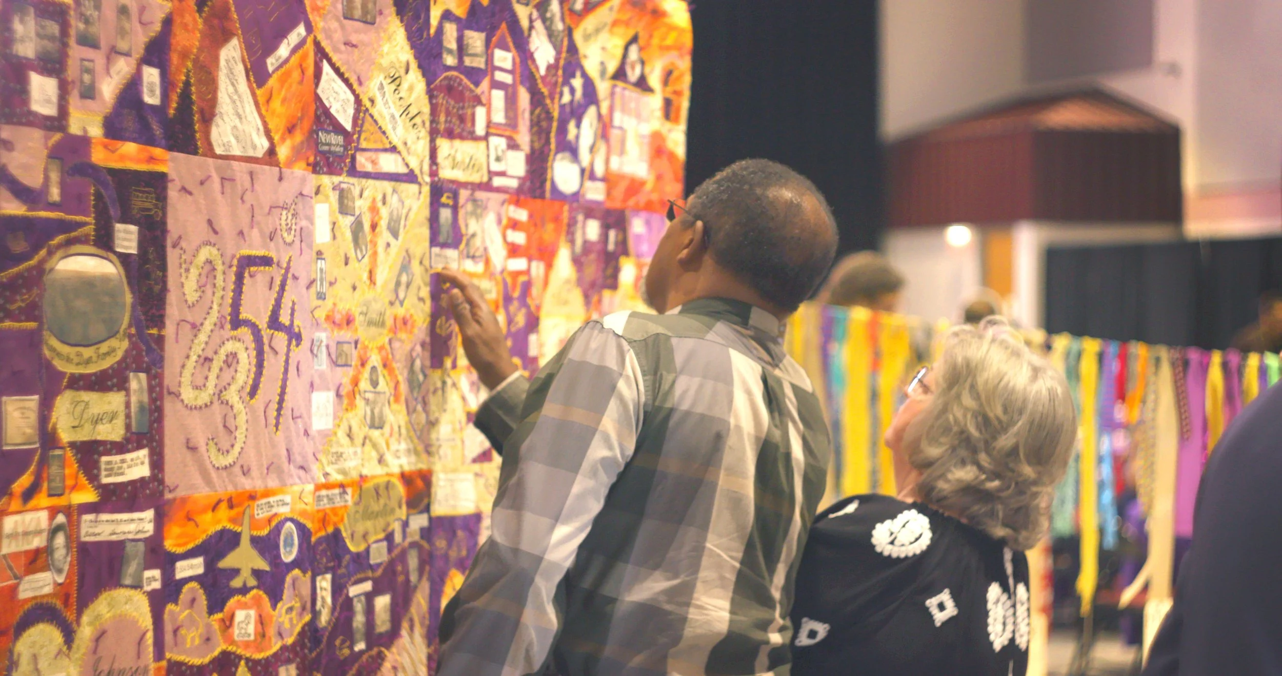 Two people, a man with dark skin and glasses, and an older woman with gray hair, are looking at a colorful textile art display.