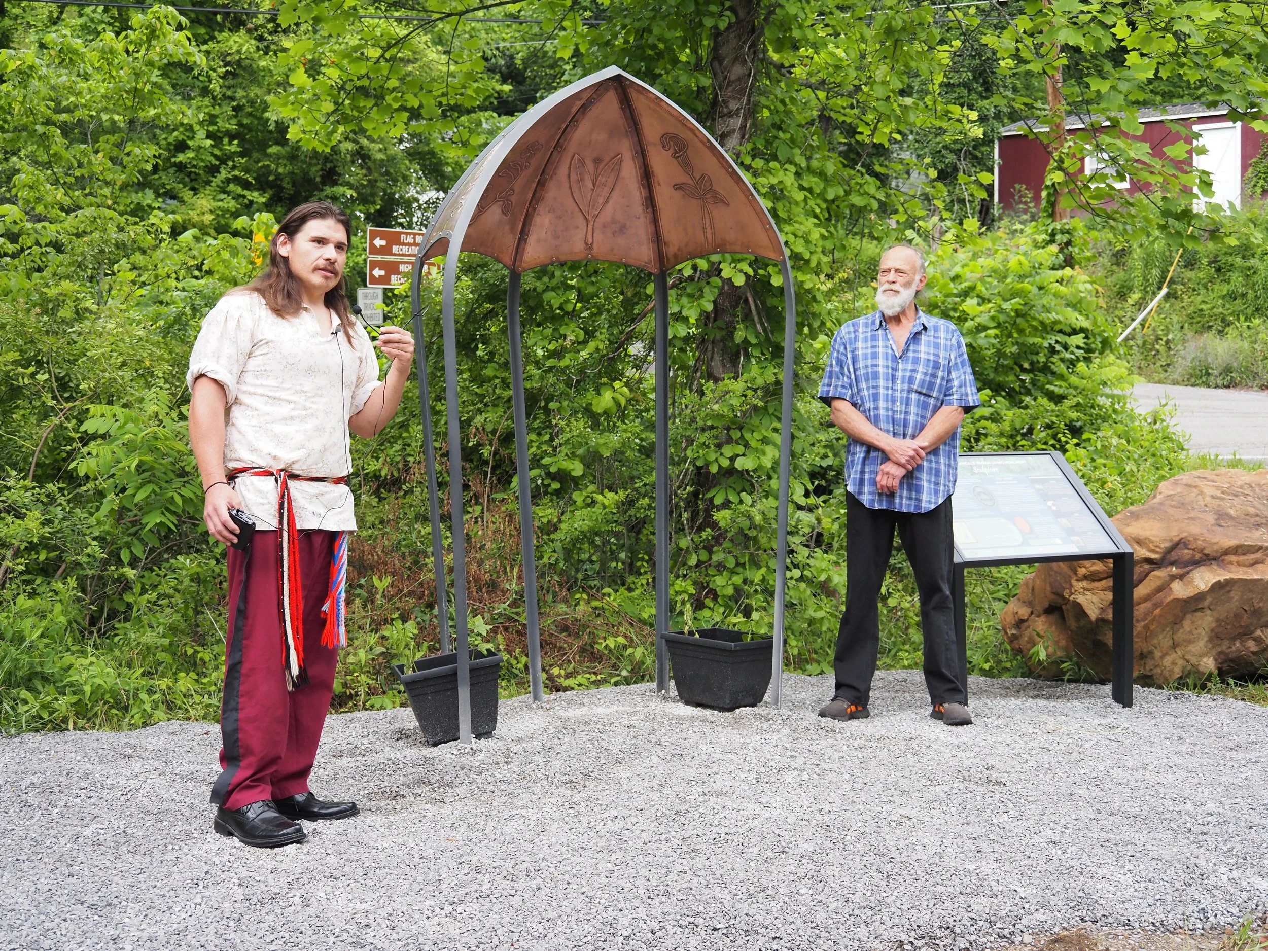 Two men standing outdoors near a small structure with a decorative roof, surrounded by green trees and bushes.