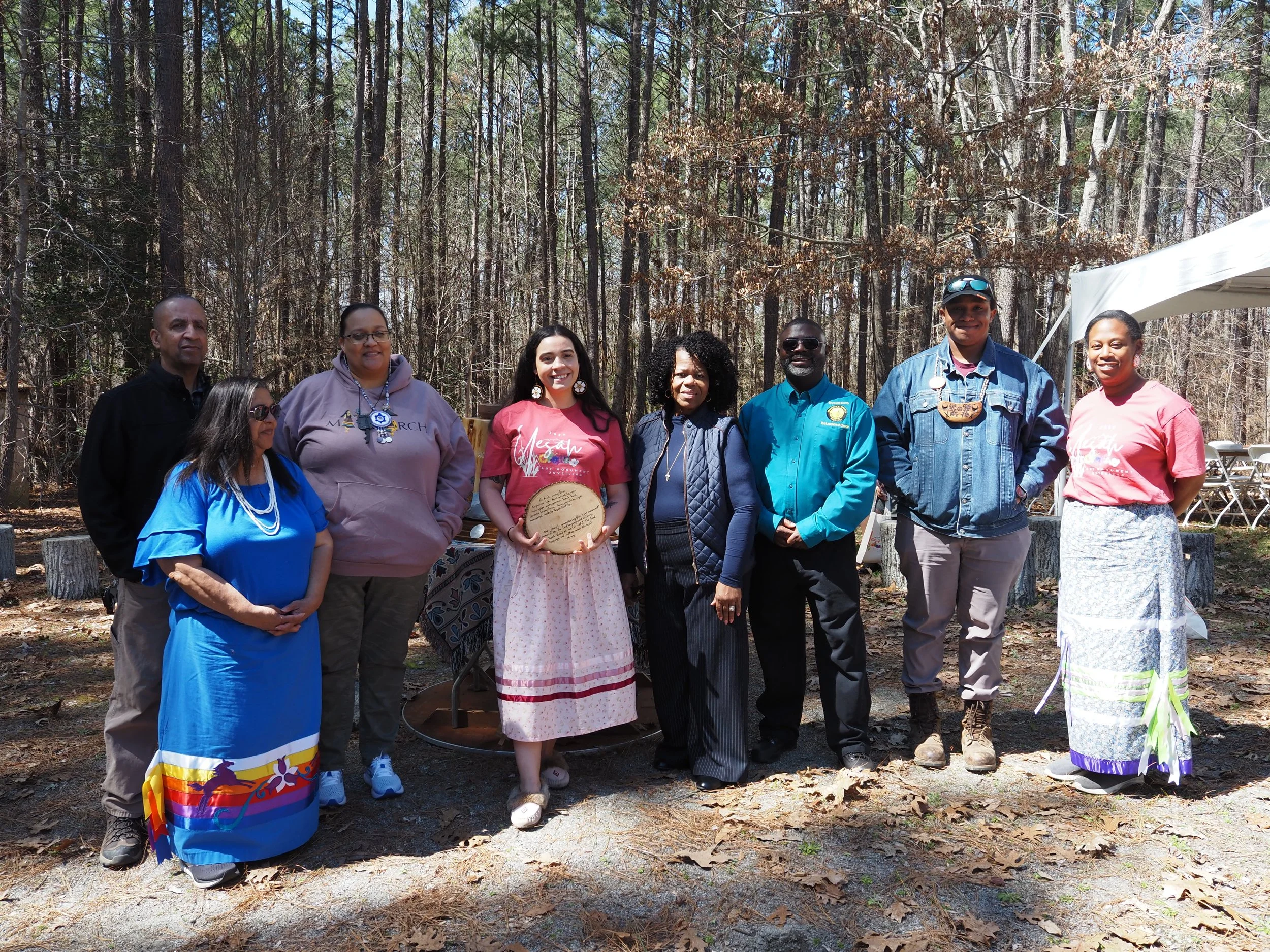 Group of nine people standing outdoors in a wooded area, some holding plaques, during daytime.