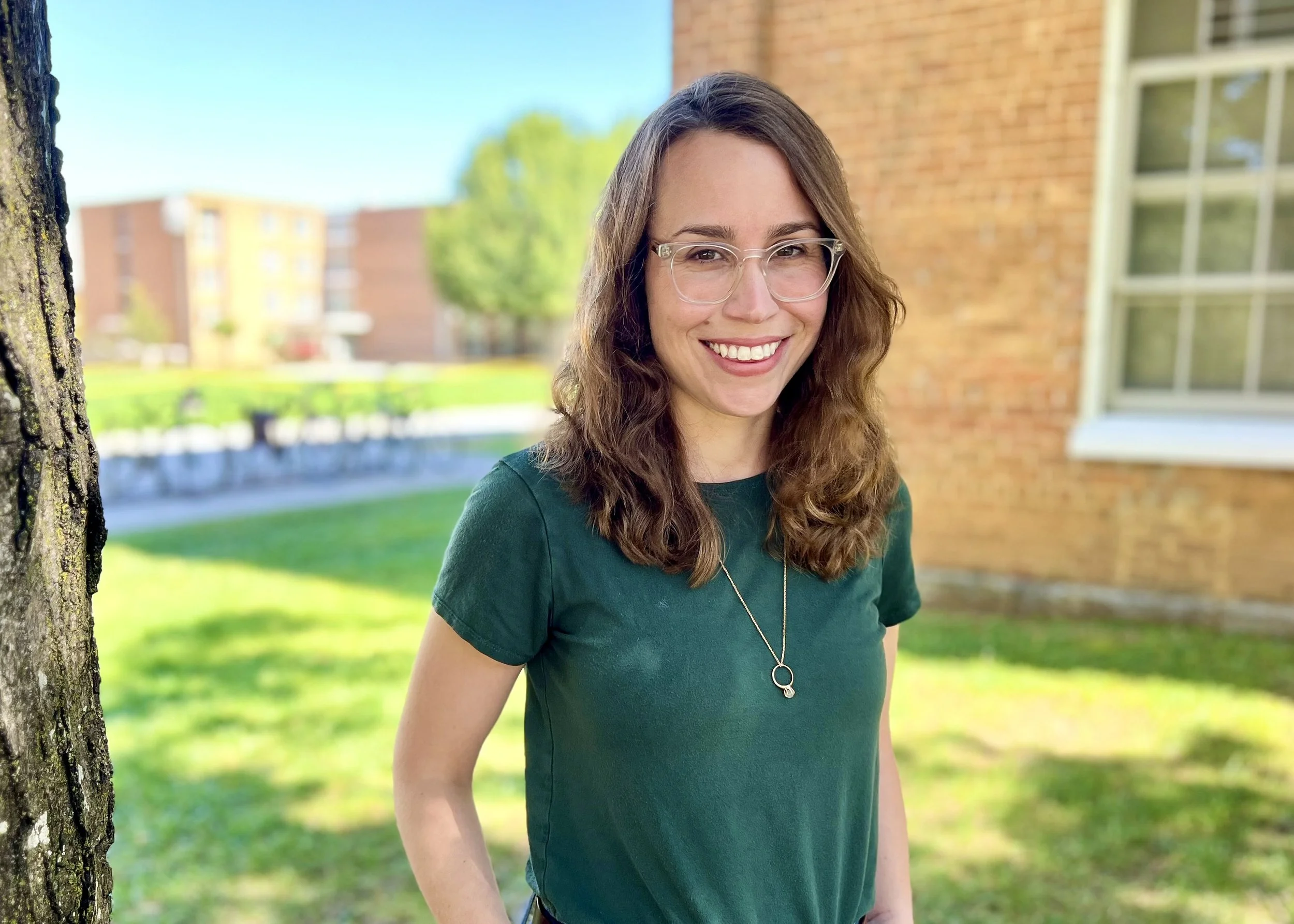 A young woman in a navy blazer and white shirt standing with her arms crossed against a brick wall, smiling.