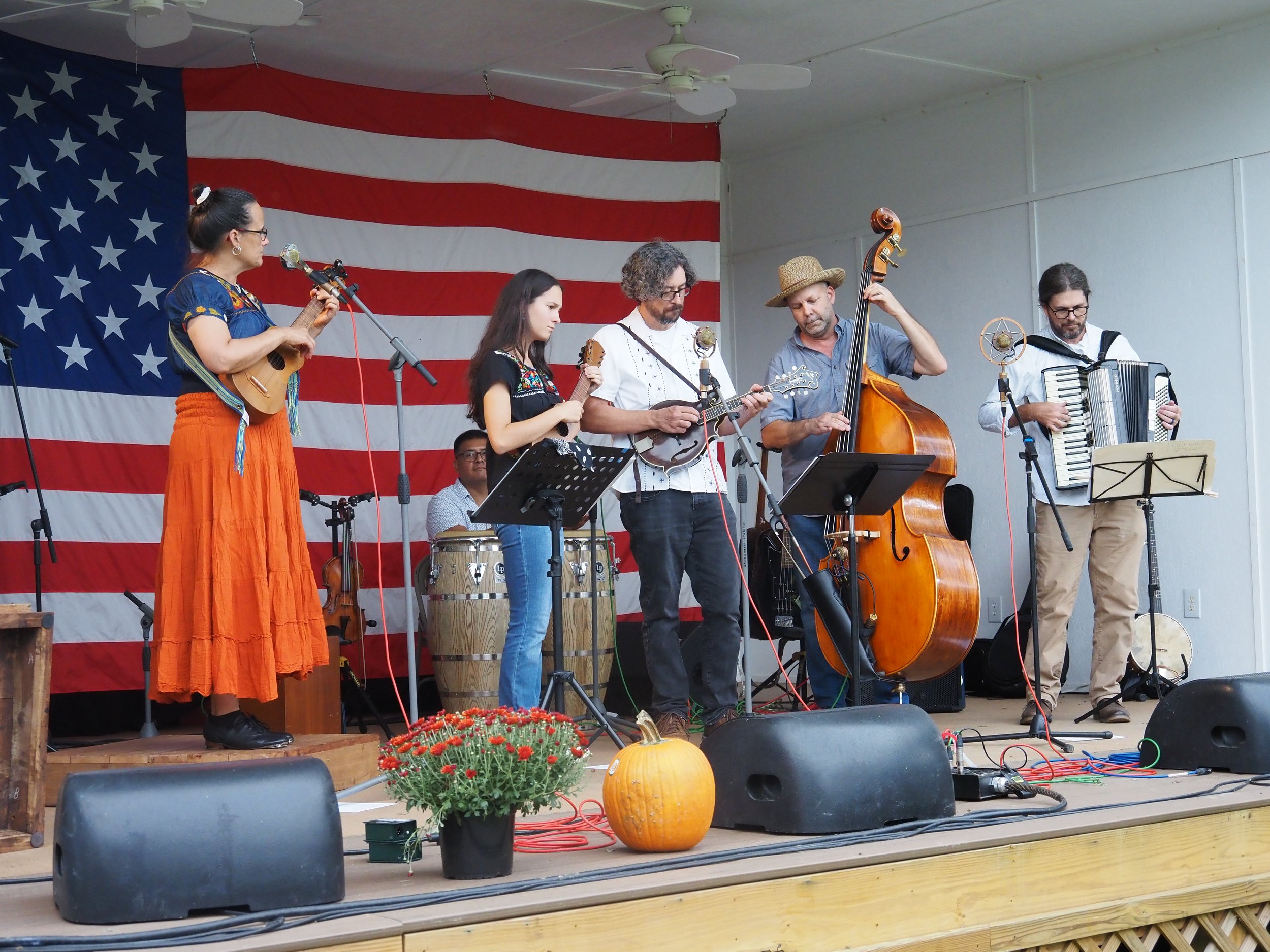 A band of six musicians performing on stage, with a large American flag as the backdrop. 