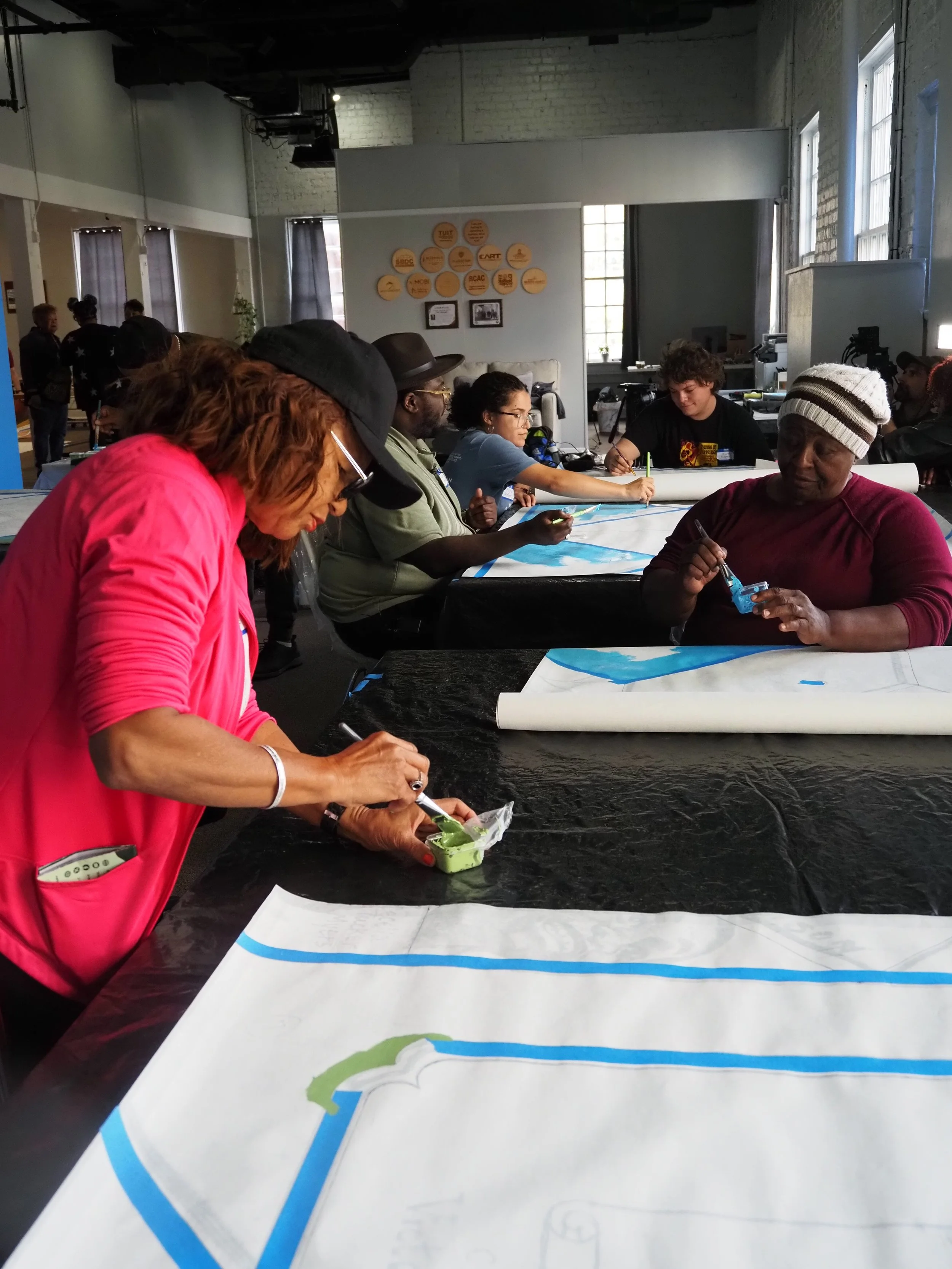 Group of diverse people painting large blue and green mural on tables in an indoor art workshop