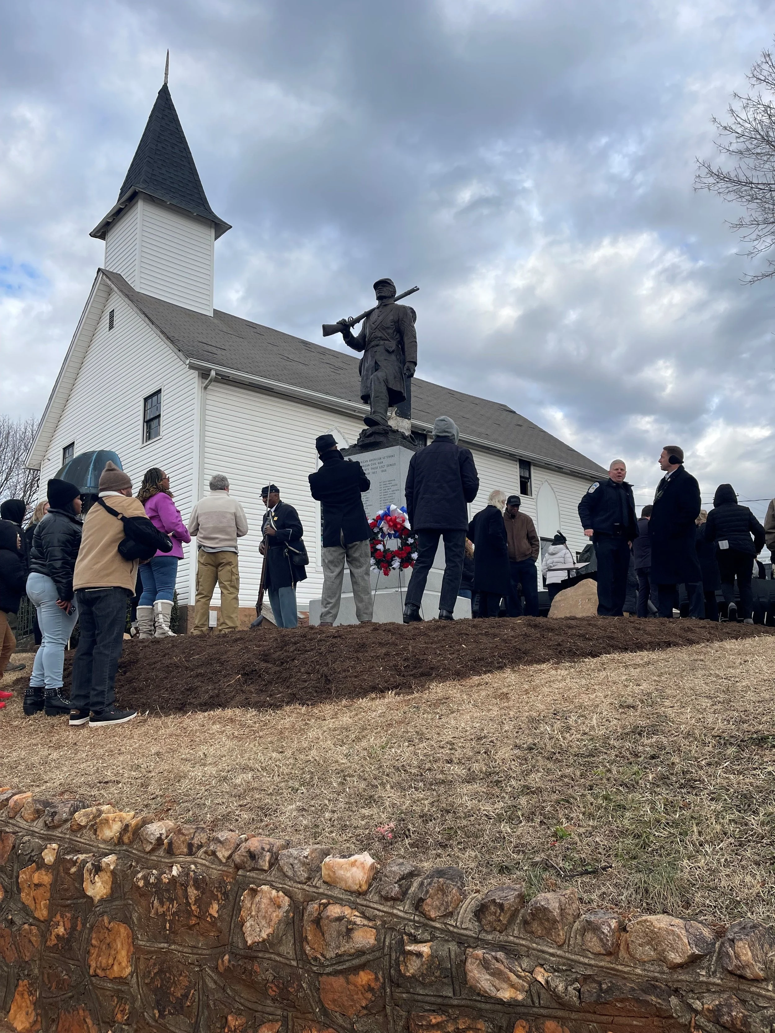 People gathered in front of a white church with a tall steeple for a statue unveiling ceremony.