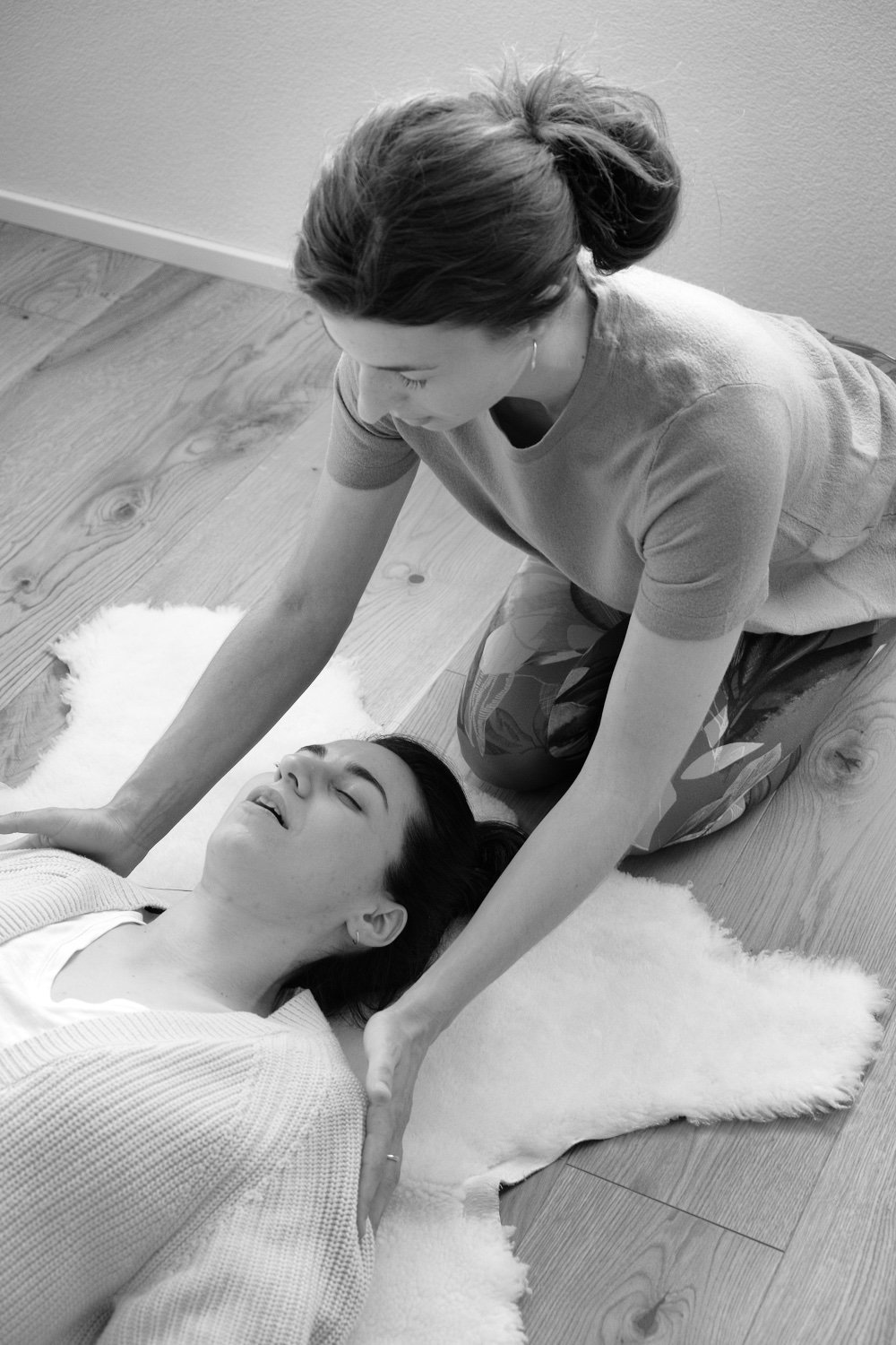 A woman receiving a head massage or chiropractic adjustment while lying on a fluffy rug, with another woman assisting on her head and neck in a home setting.