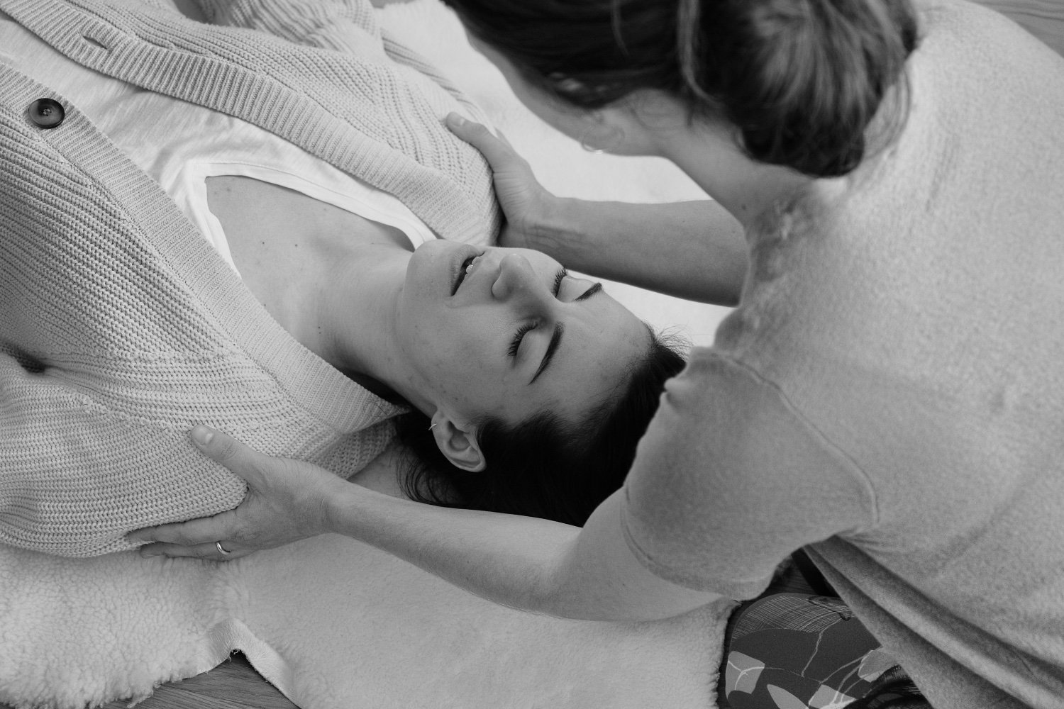 A woman with her eyes closed lying on a bed, receiving gentle care from another woman who is leaning over her, with her hands on the woman's shoulders and chest.