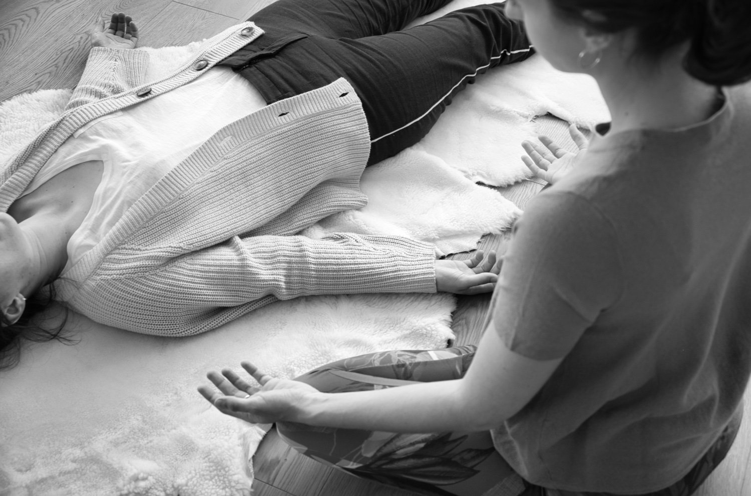 A woman lying on her back on a fluffy rug with her arms relaxed at her sides, and another woman kneeling beside her with one hand raised, engaging in conversation or possibly performing a medical or wellness procedure.