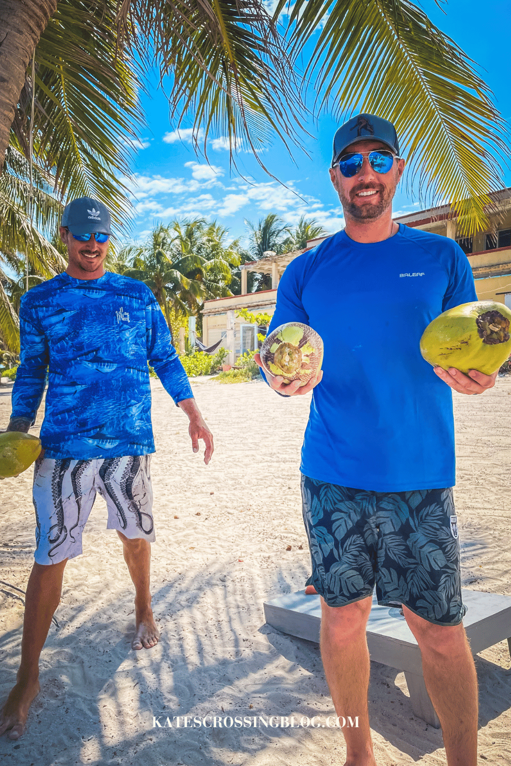 Kate's husband and another man are standing on a beach, holding up green coconuts and smiling, with palm trees behind them. 