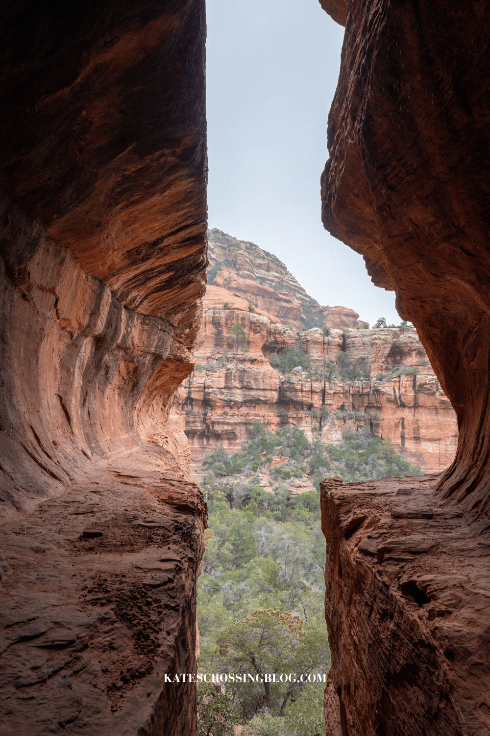 View from inside subway cave, framed by towering red rock walls, looking out towards the stunning Sedona landscape with vibrant red rock formations and green trees.
