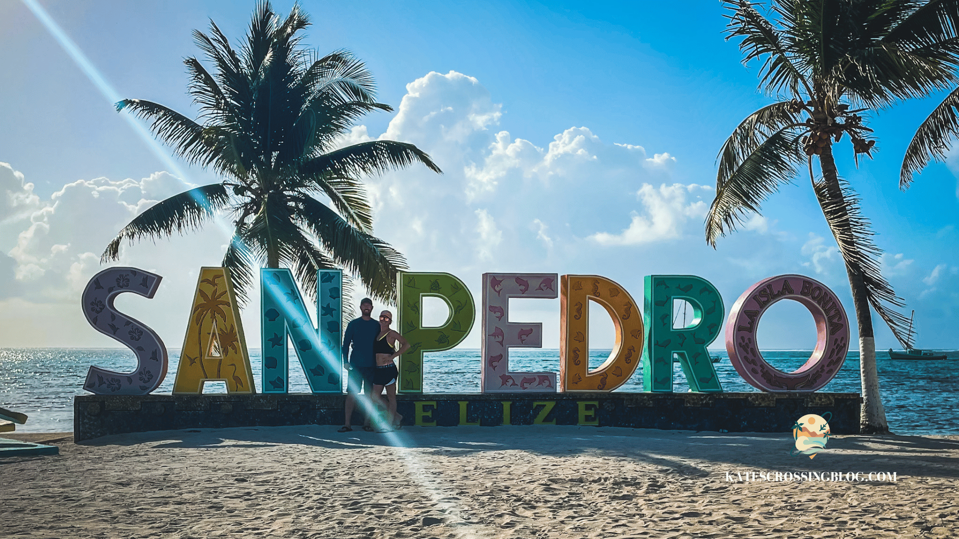 Kate and her husband posing on a beach with a colorful "San Pedro" sign with palm trees and the ocean in the background. 