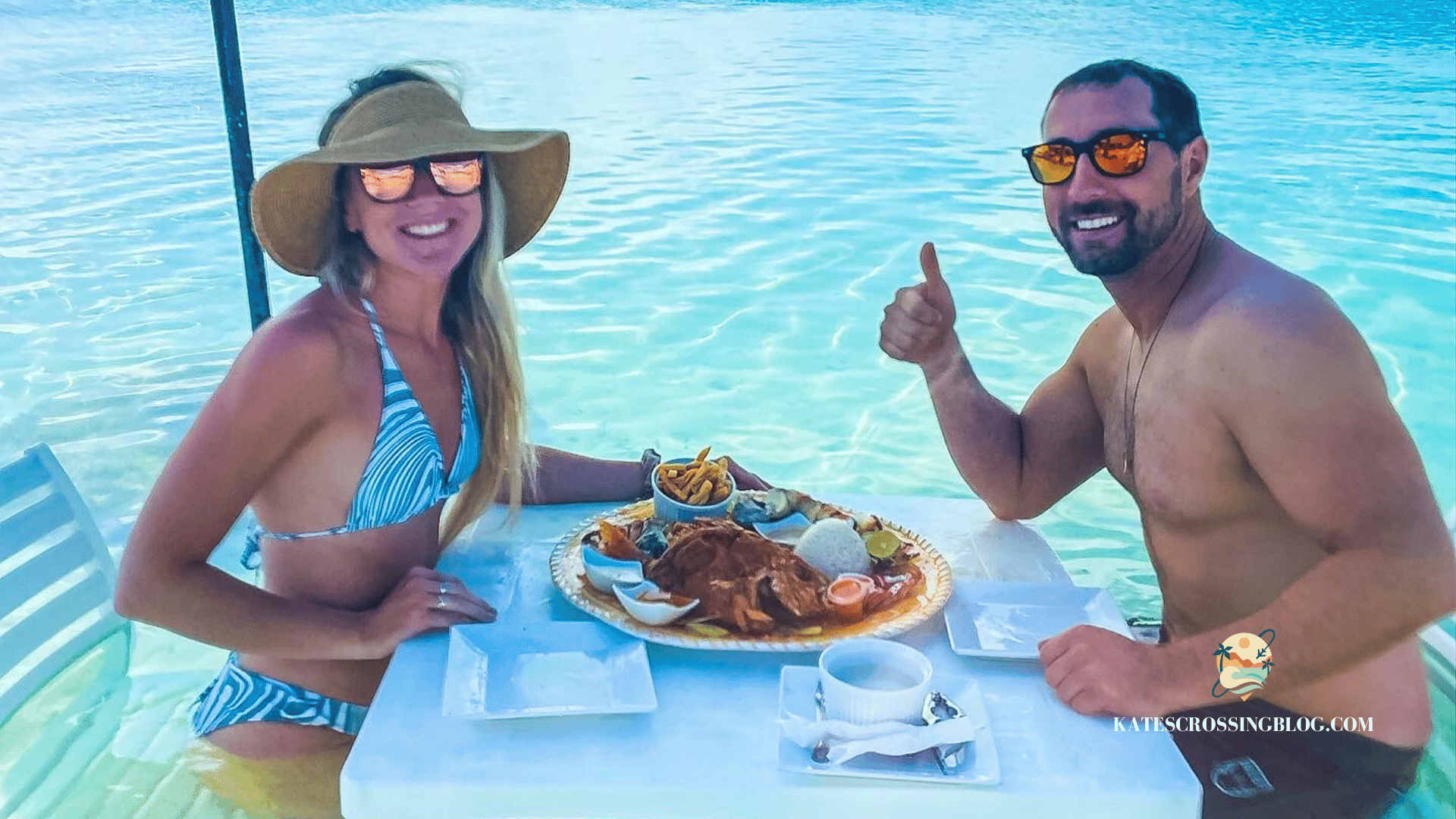 Kate and her husband sitting at a picnic table out in the turquoise water off the shore of Secret Beach, eating a seafood platter, shaded by a yellow beach umbrella. 