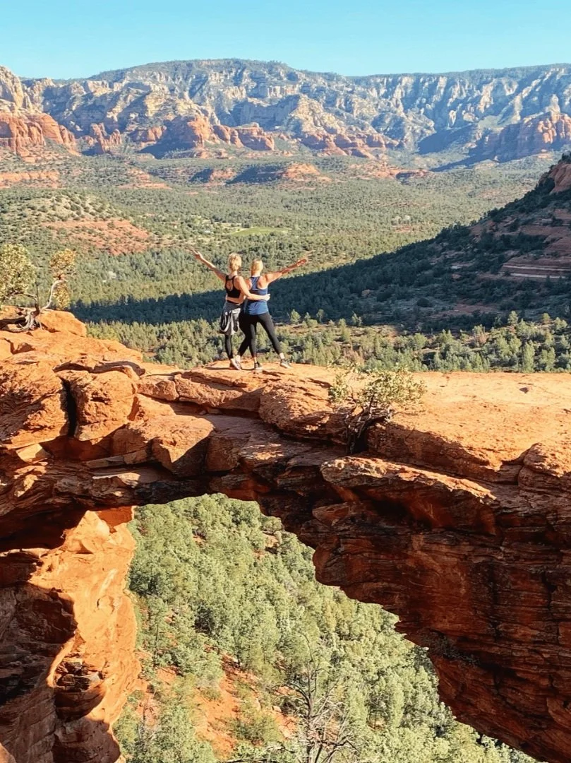 Kate and a friend standing on Devil's Bridge, a popular hike in Sedona, with a stunning backdrop of red rock formations and desert valley.