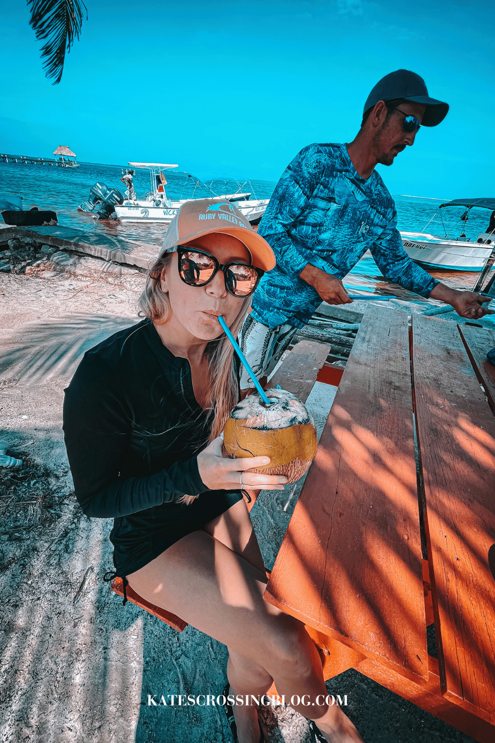 Kate sitting at a picnic table, drinking a fresh coconut through a blue straw, with boats and the ocean behind her. 