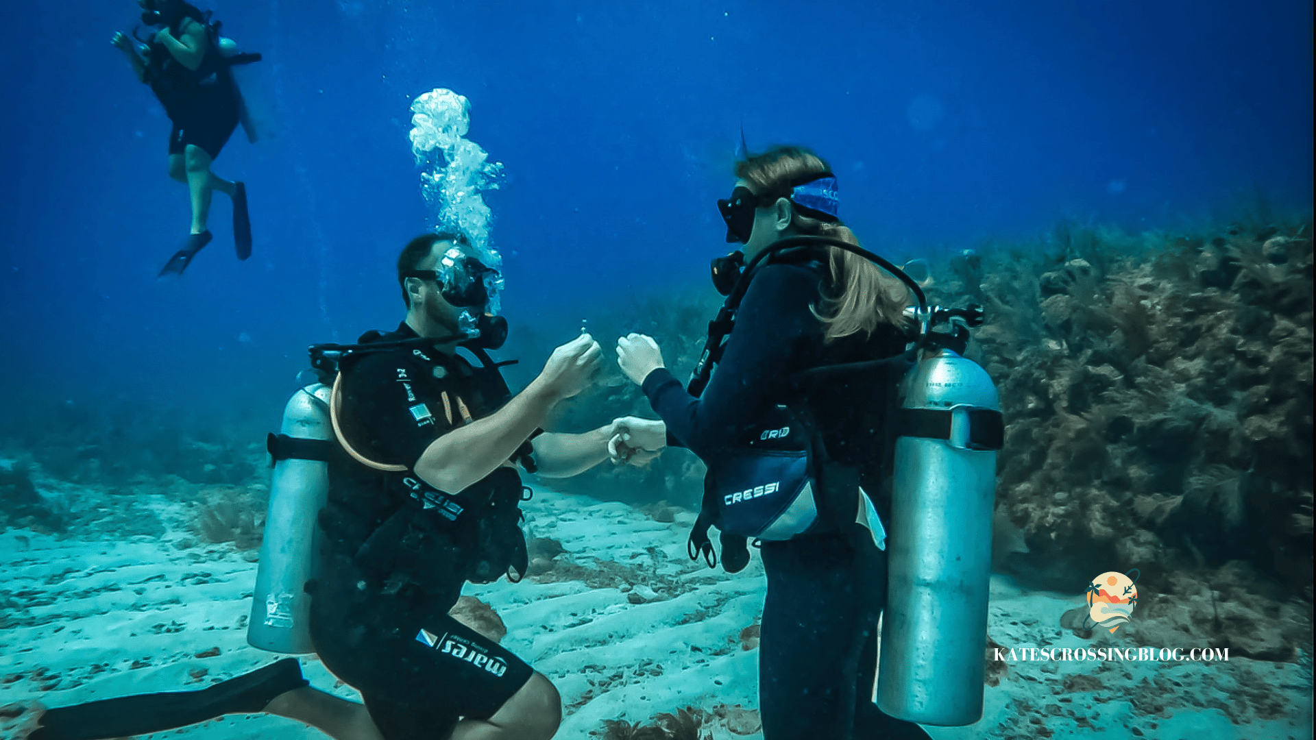 Kate getting proposed to on the ocean floor while scuba diving with man down on one knee holding out a ring, and coral formations in the background. 
