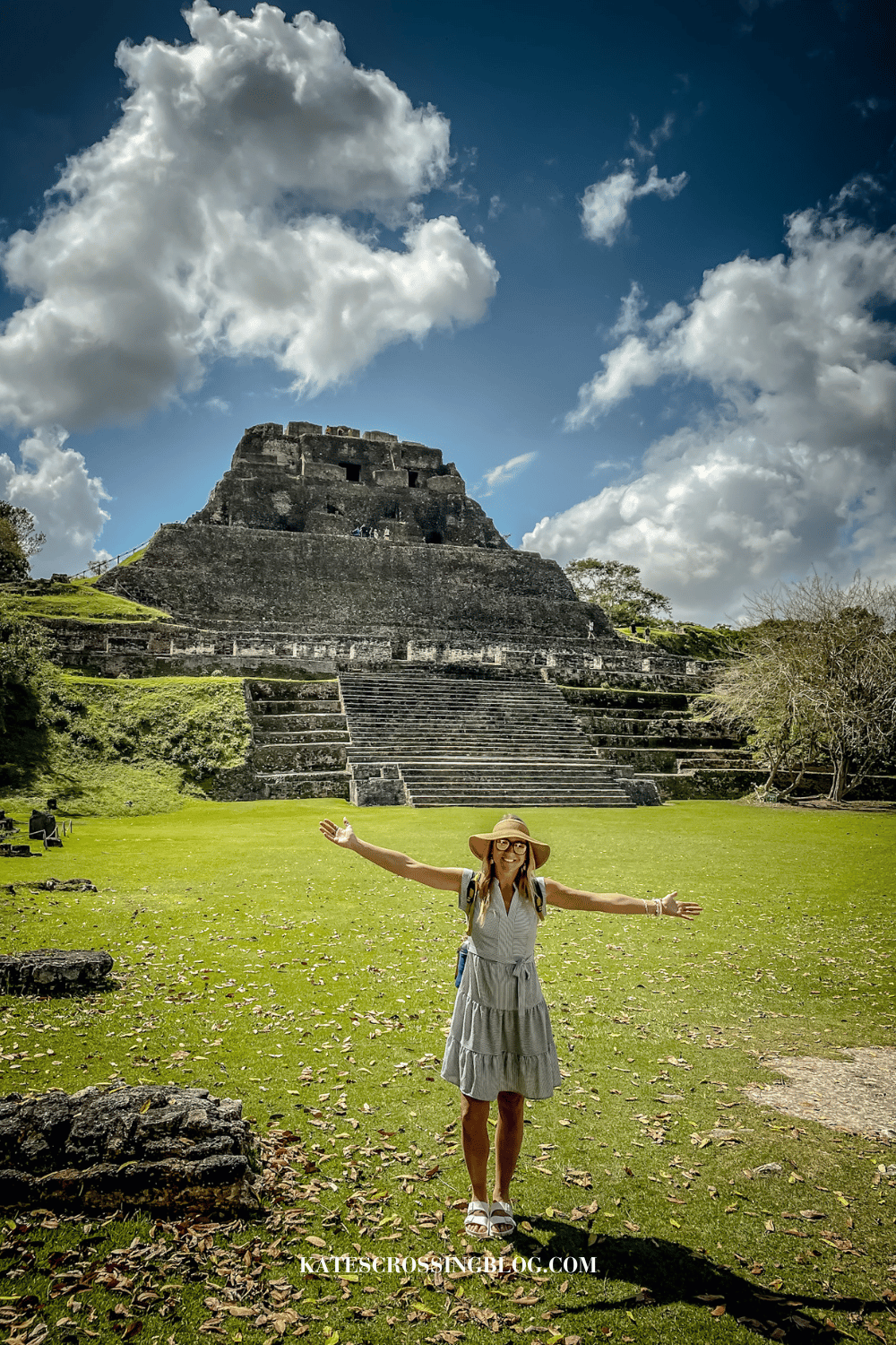 Kate standing with arms reached out in front of the Mayan Temple Xunantunich, with green grass and jungle trees around the temple.