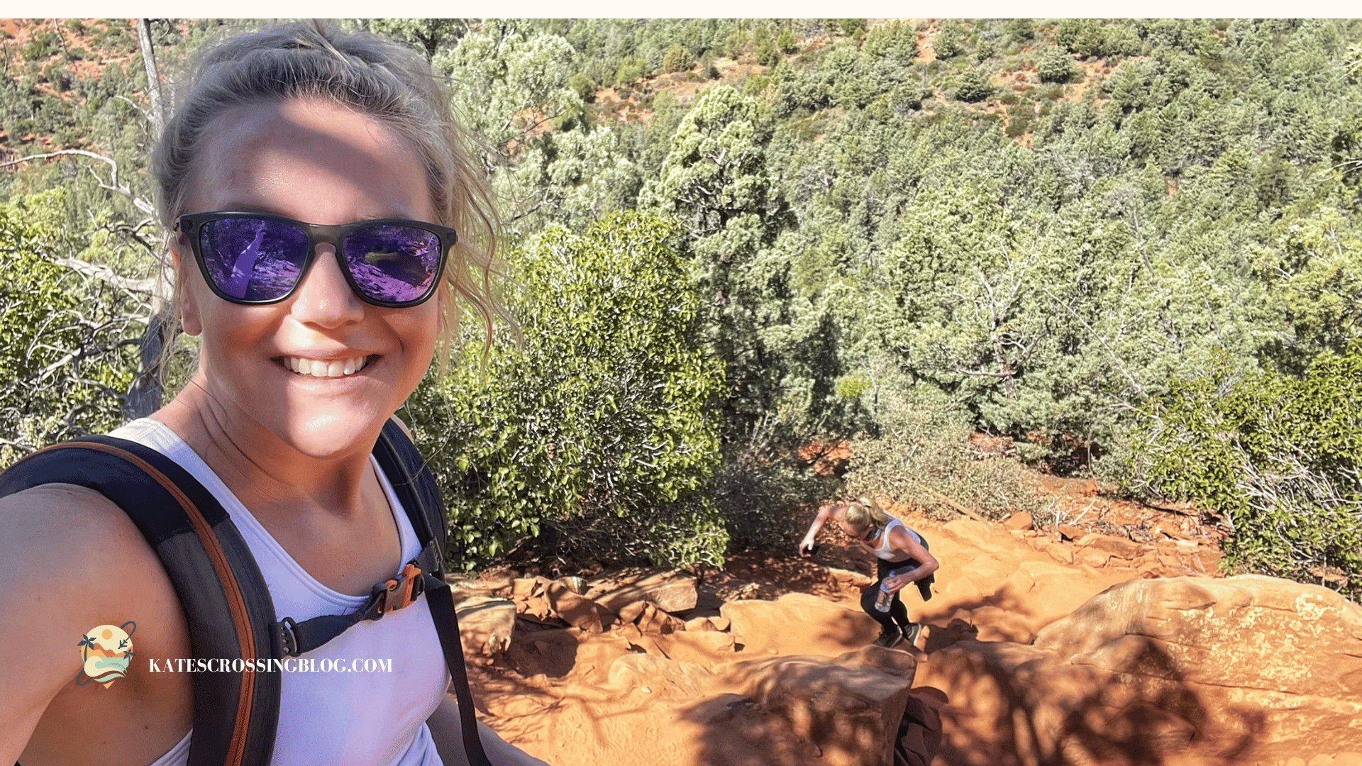 Kate smiling for a selfie along the Soldier Pass Trail in Sedona, with a friend hiking up the rocky path in the background, surrounded by desert trees.