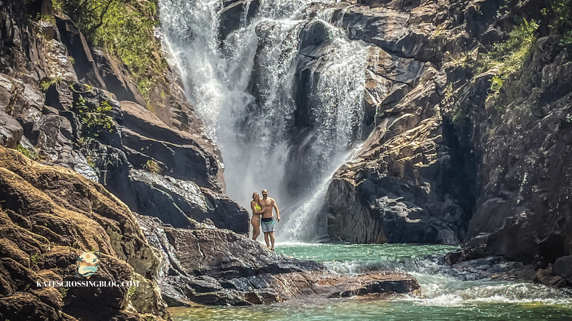 Kate and her husband are standing on rocks below the Big Rock Falls waterfall, with green vegetation growing on the slippery rocks. 