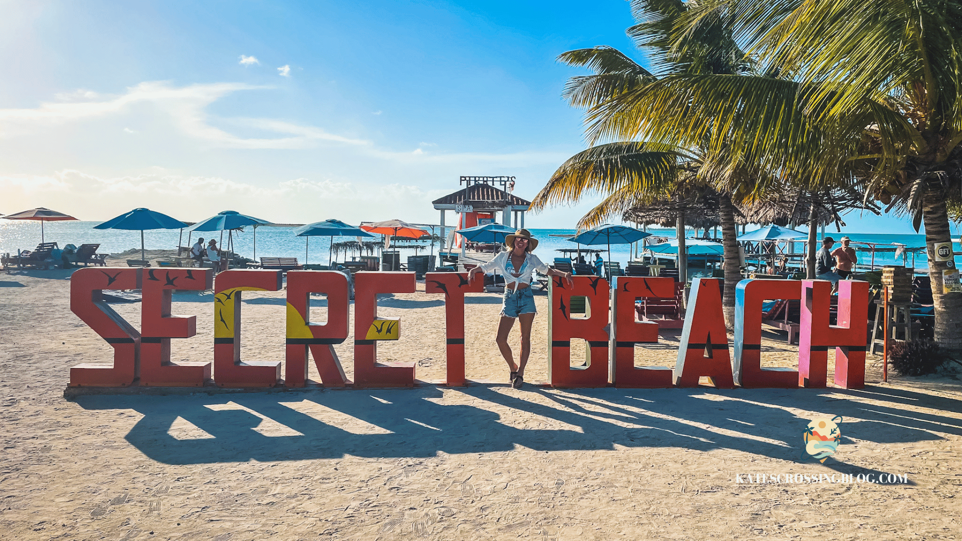 Kate standing next to a orange "Secred Beach" sign with beach umbrellas, palm trees and the ocean in the background. 