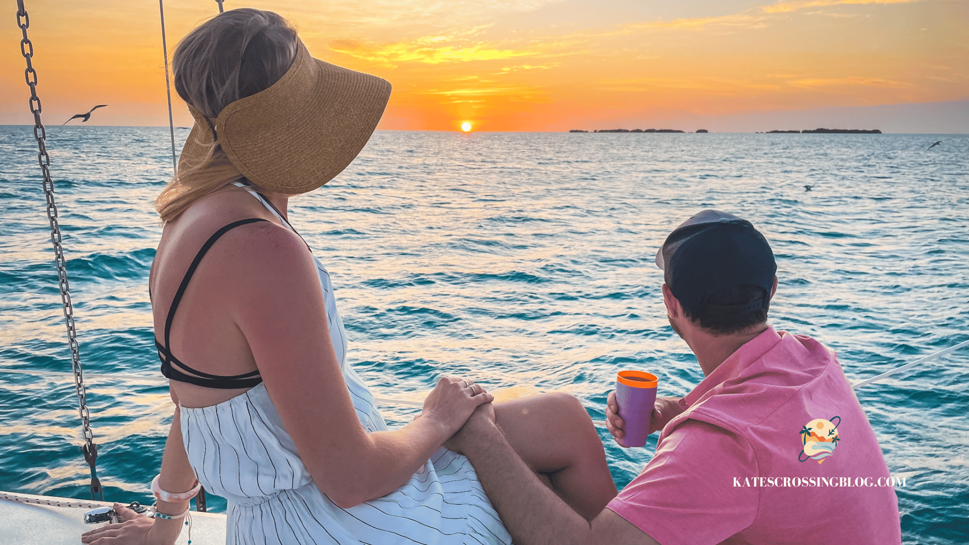 Kate and her husband are sitting on the side of a sailboat, looking out at a sunset over the water, holding hands. 