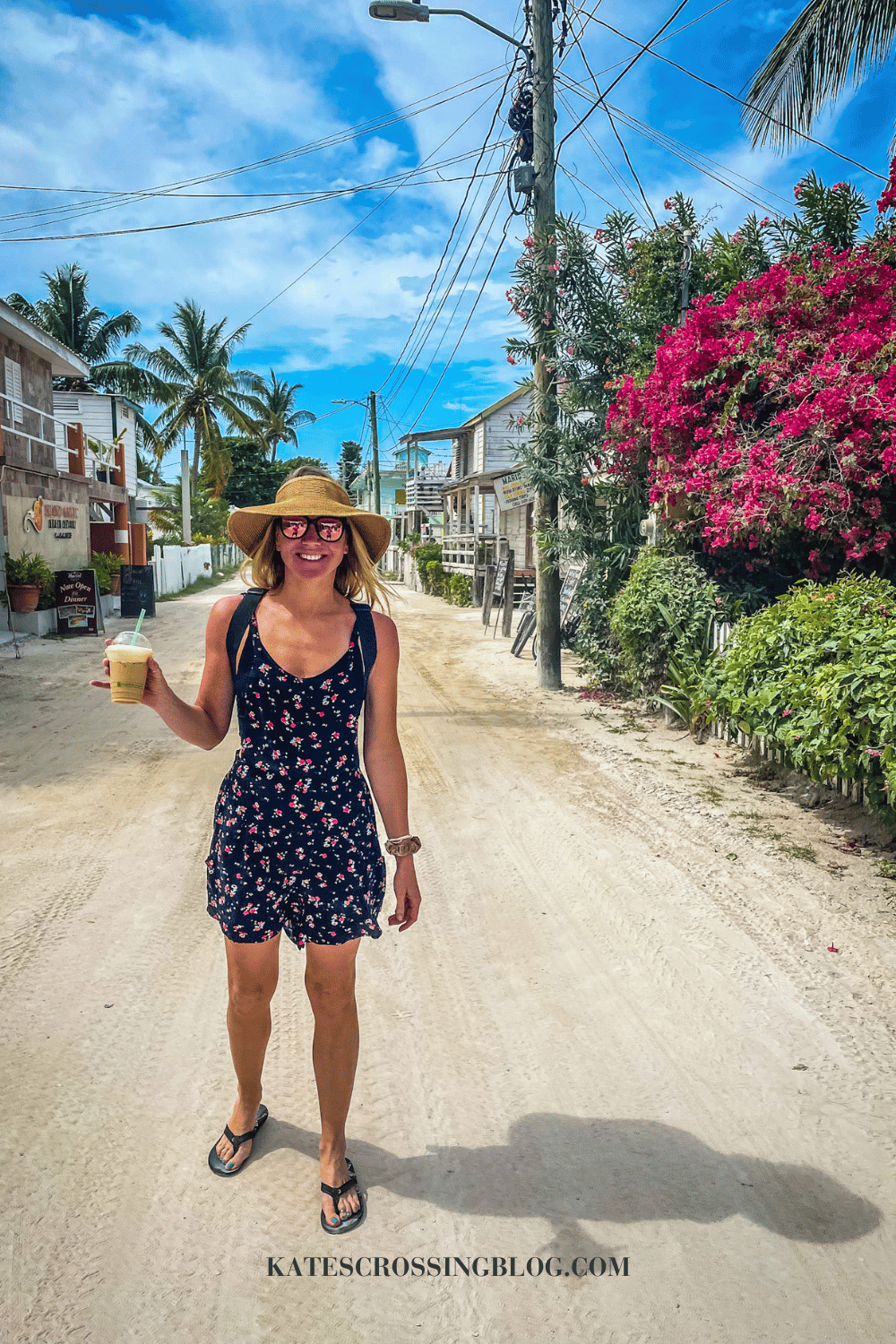 Kate walks a sandy street in Caye Caulker, lined with stores, palm trees, and colorful tropical flowers, holding a coffee in a floral sundress and sunhat. 