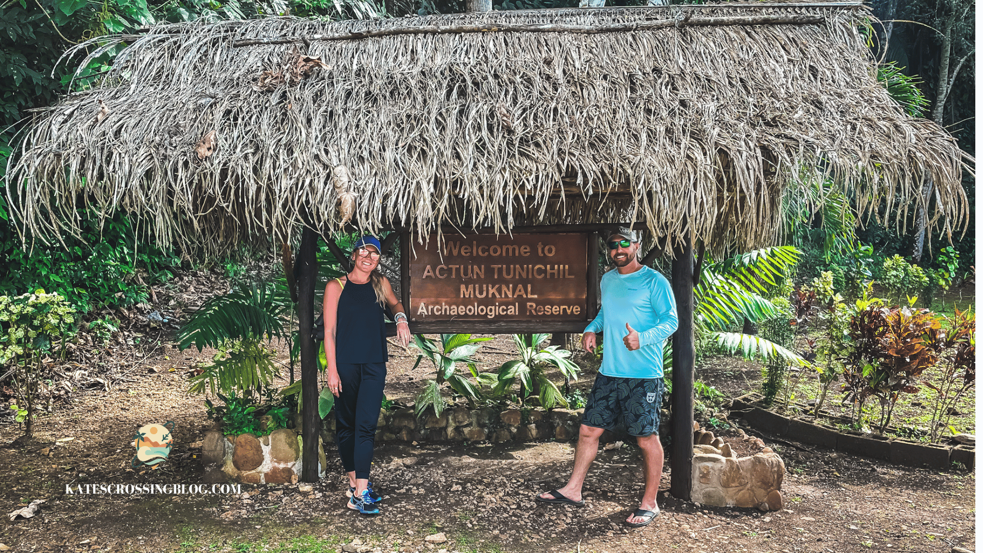 Kate and her husband posing next to the "Welcome to the ATM cave" sign, covered by a thatched roof with jungle in the background.