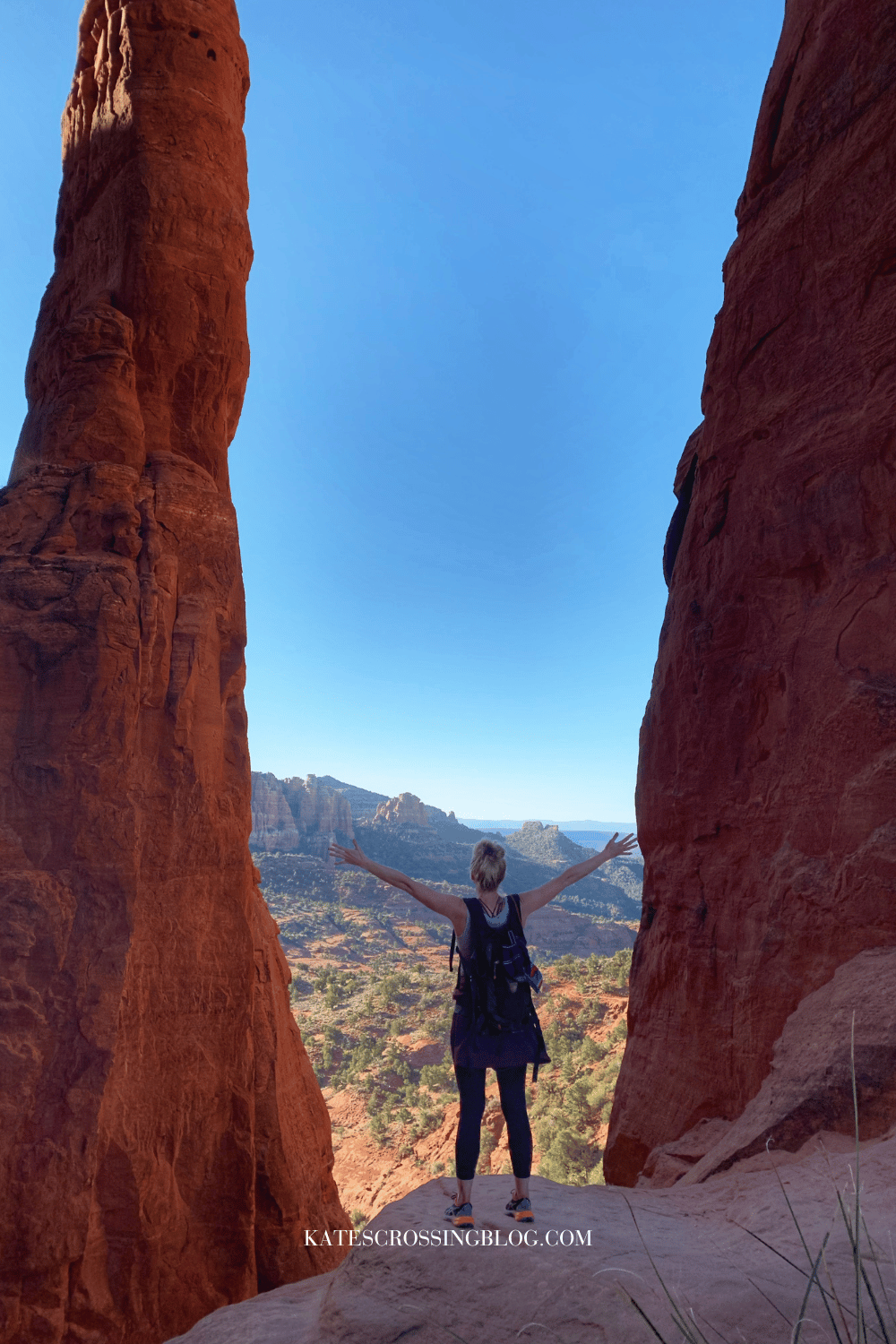 Kate stands between two tall red rock formations, overlooking a breathtaking view of Sedona's landscape with arms outstretched, embracing the natural beauty.