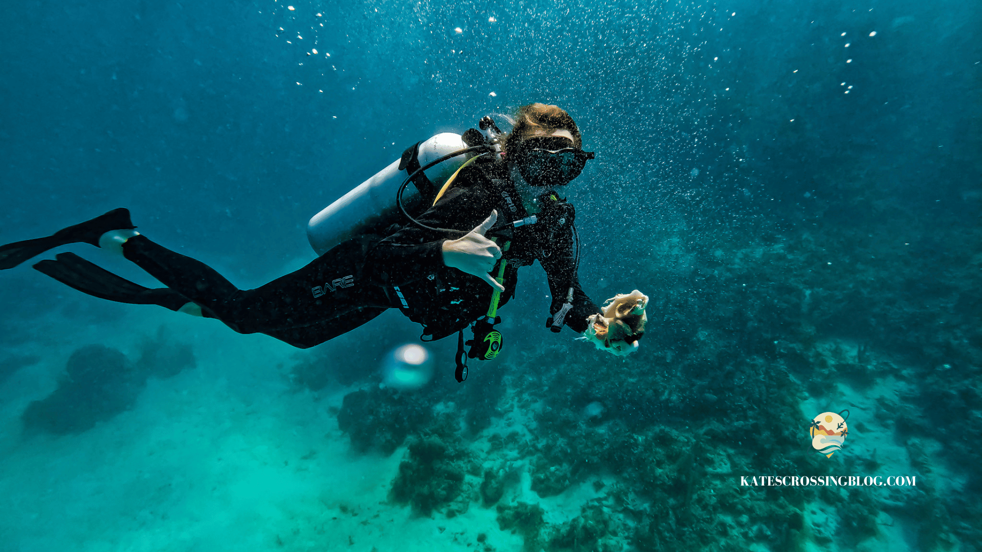 Kate scuba diving in turquoise water, giving a thumbs up sign while holding a conch shell.