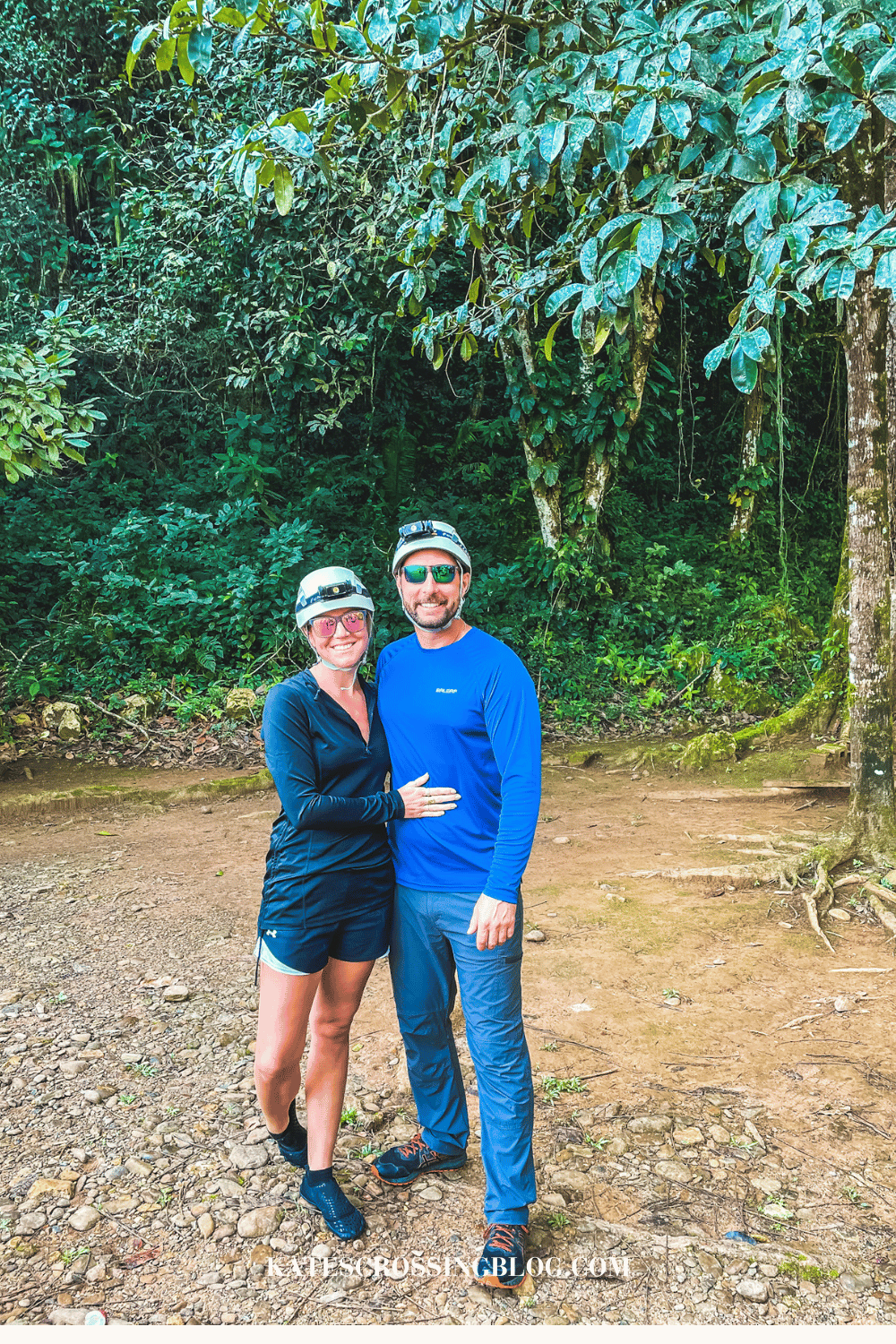 Kate and her husband standing together with helmets, headlamps and hiking clothes, with the jungle behind them.