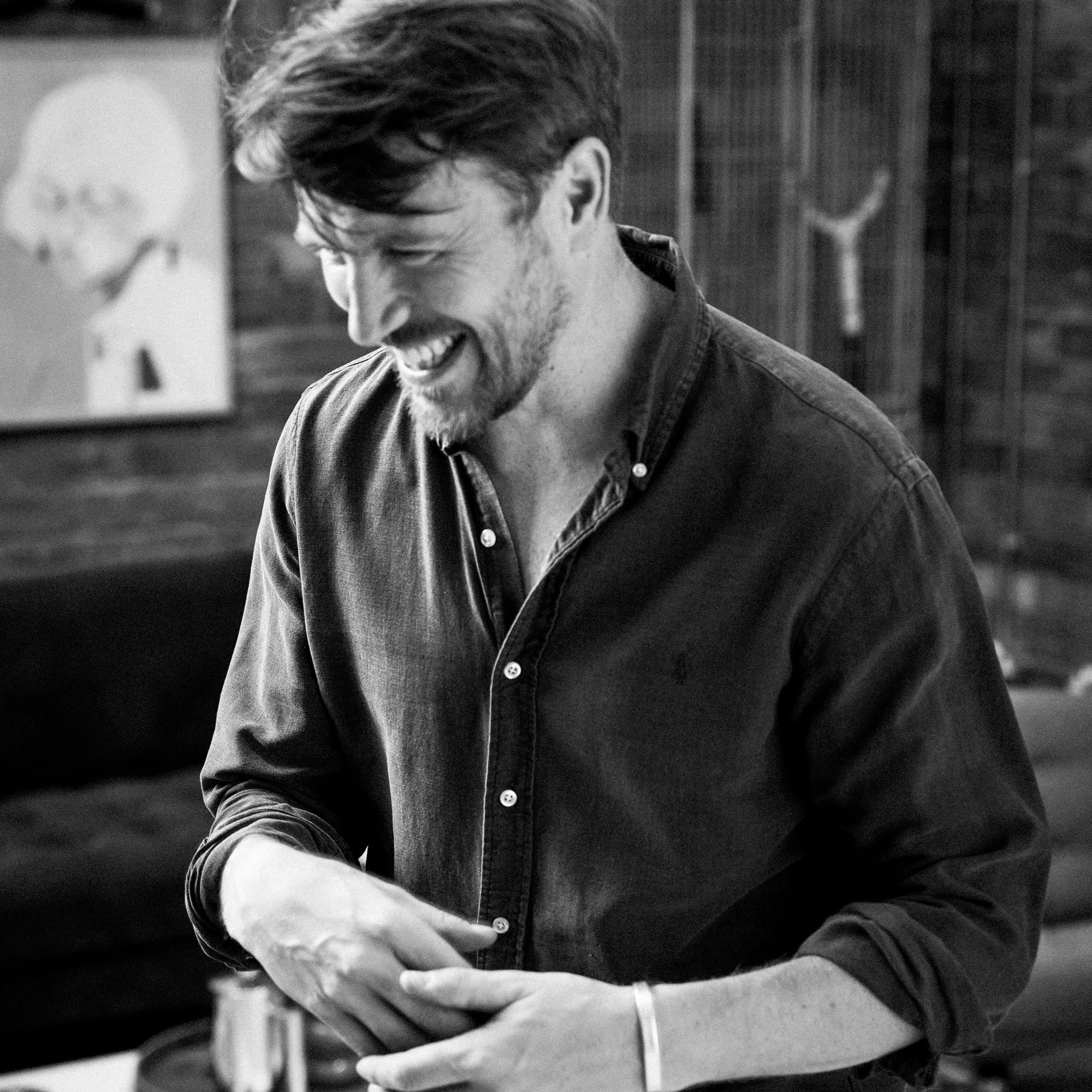 A young man and a beard smiling and looking down, wearing a denim shirt, with a blurred background that appears to be an office or creative workspace.