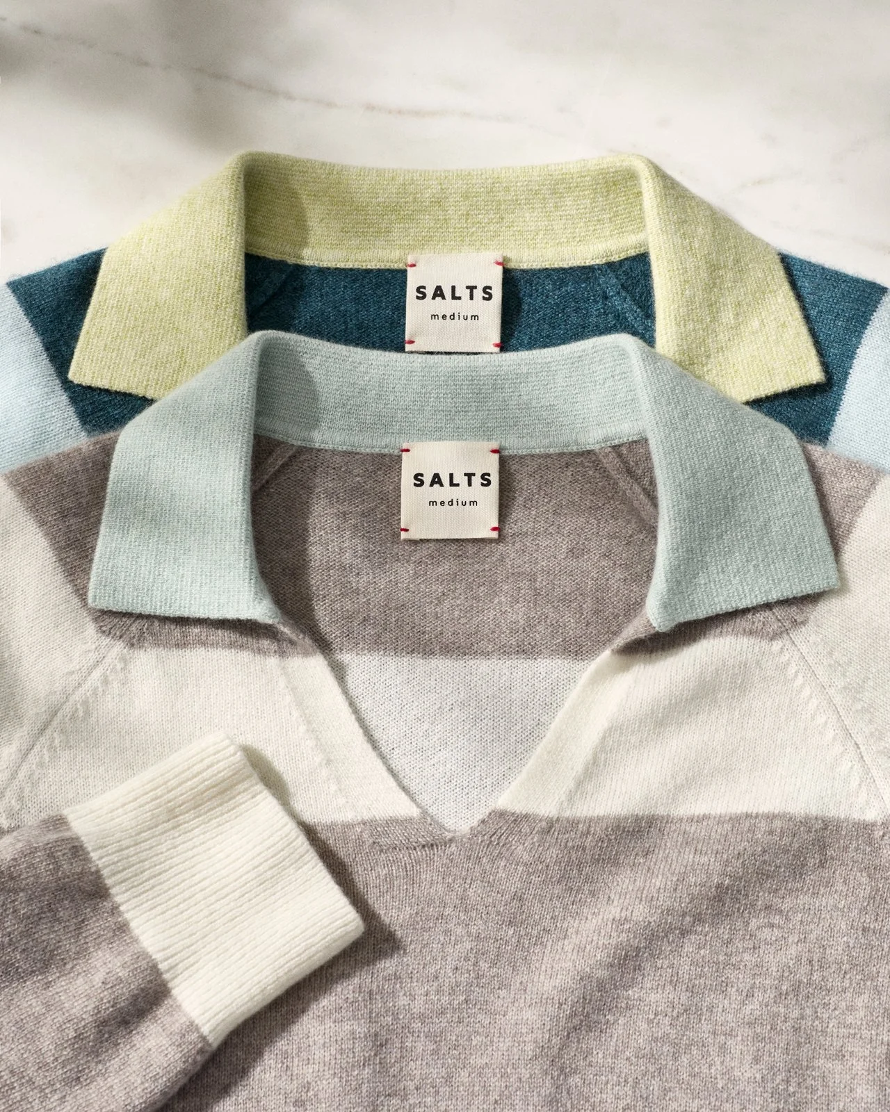 Close-up of three folded striped sweaters with collars, labeled 'SALTS medium.' The sweaters are in light pastel colors: green, blue, and gray with beige and white stripes.