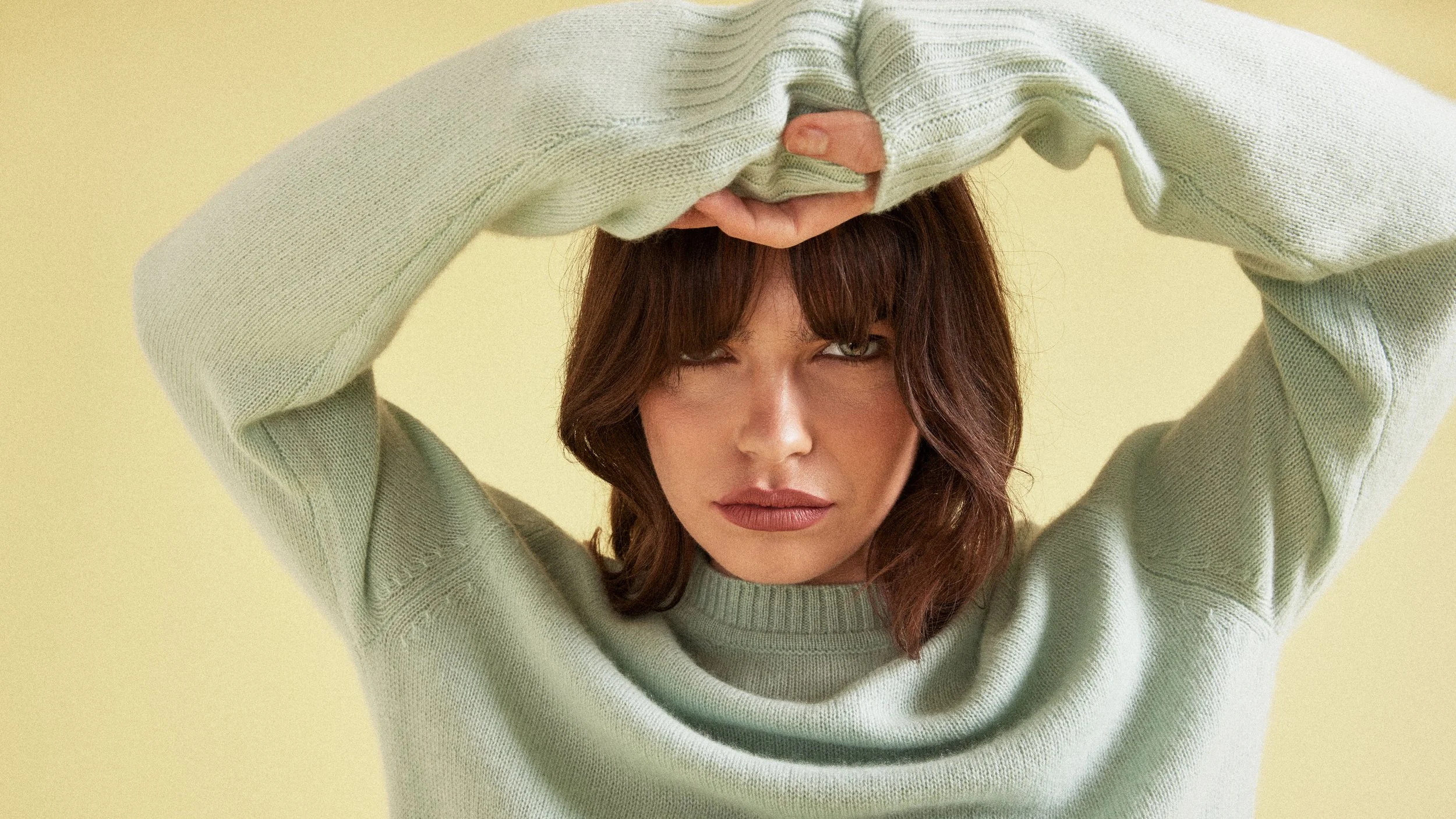A woman with brown hair and light makeup, wearing a green sweater, poses with her arms raised and one hand on her forehead, against a yellow background.