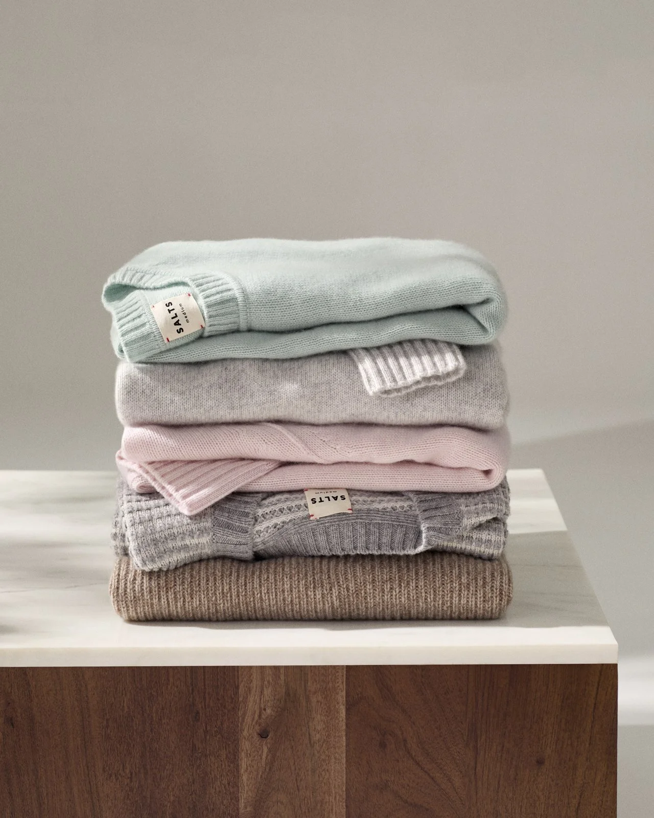 A neatly folded stack of five sweaters in pastel colors on a white surface with a plain gray background.