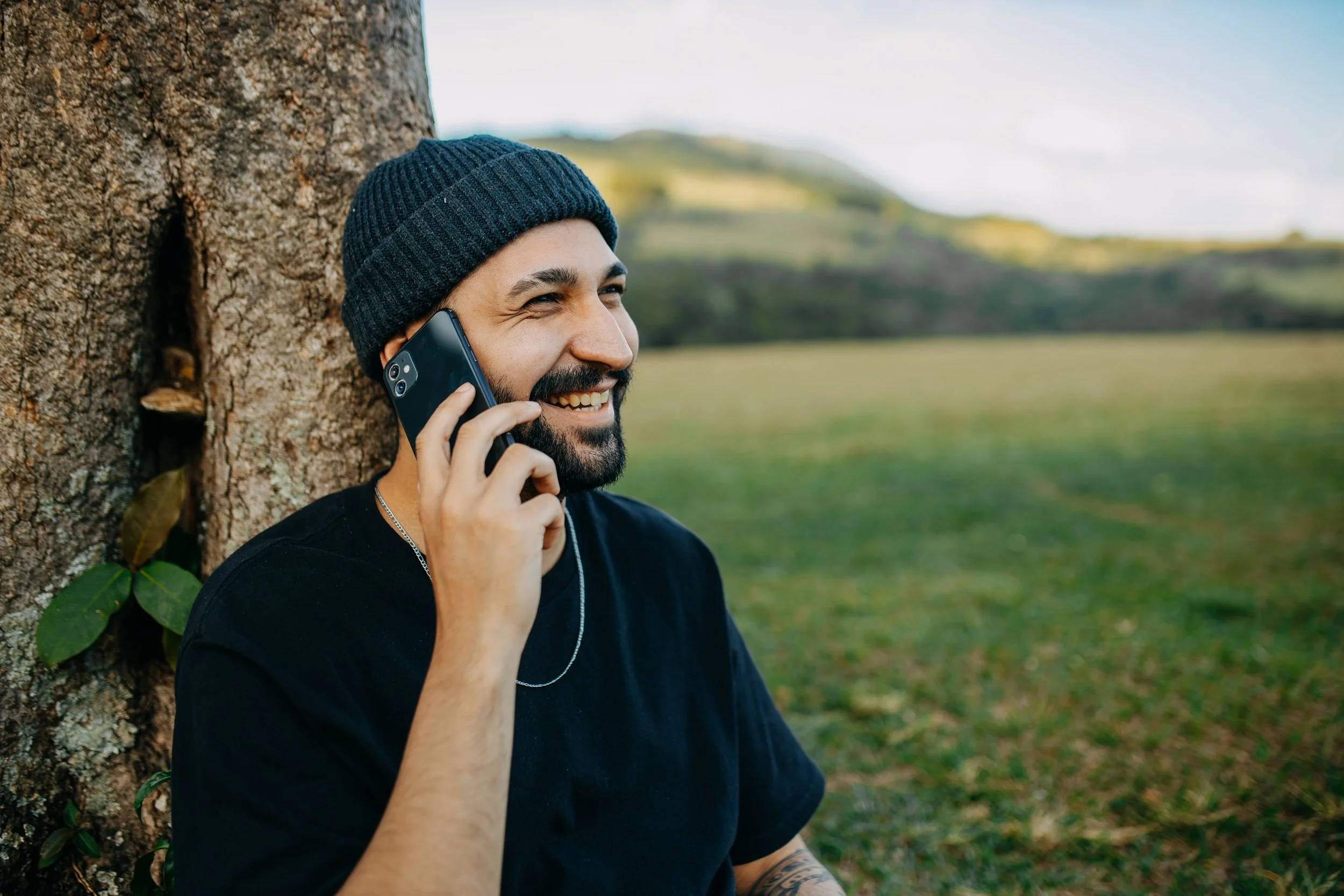 A man leaning with his back against a tree in the park, on the phone smiling