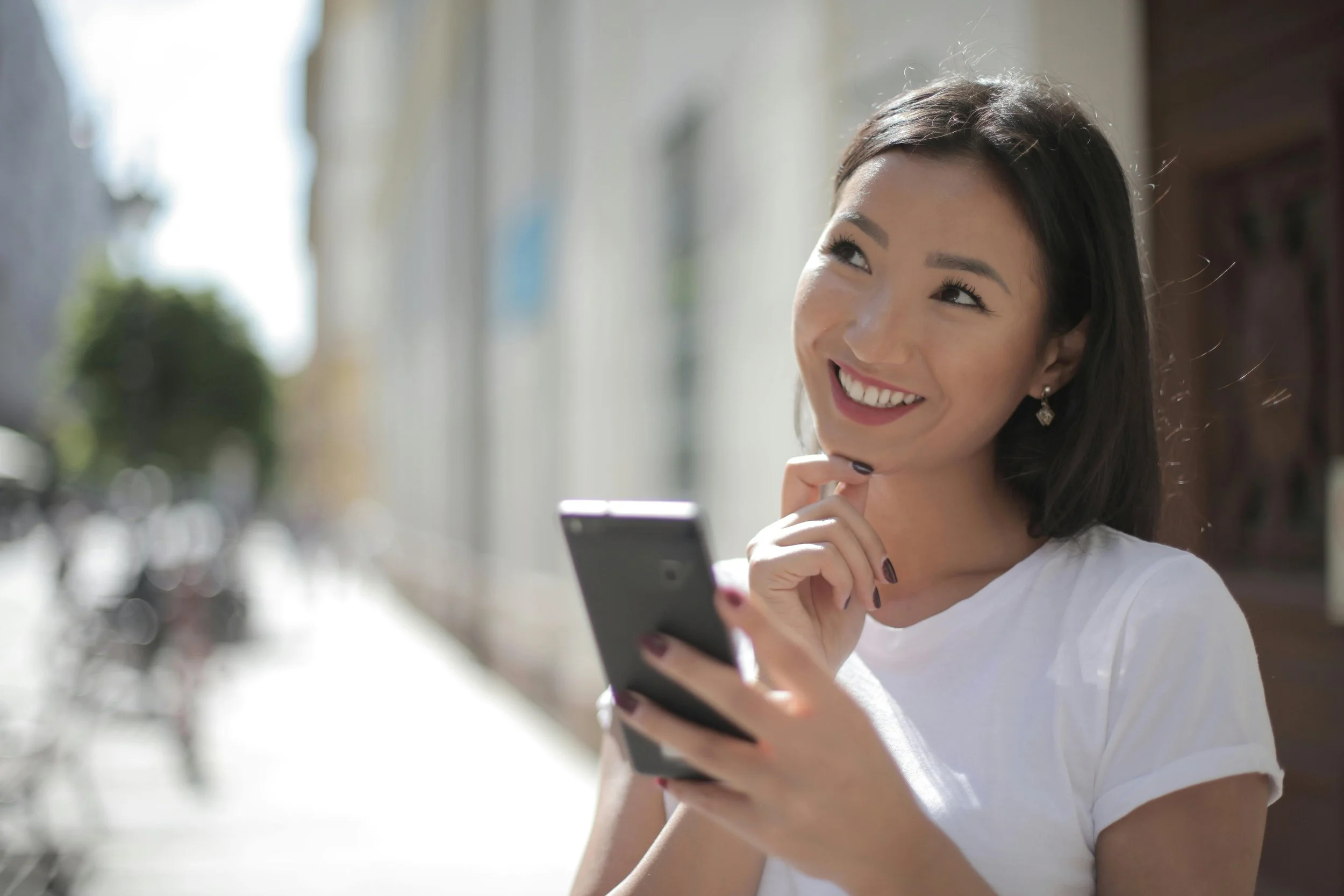 Women in white shirt holding black phone to take a selfie, while posing with her right hand under her chin and looking upward to appear to be thinking