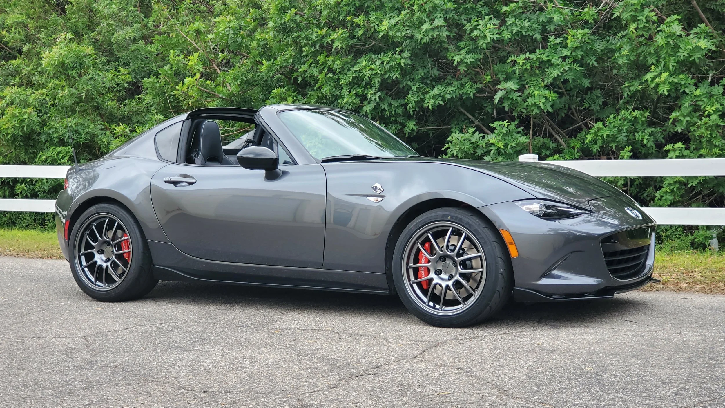 A gray BMW convertible sports car parked on the side of a paved road with a white guardrail and green bushes in the background.