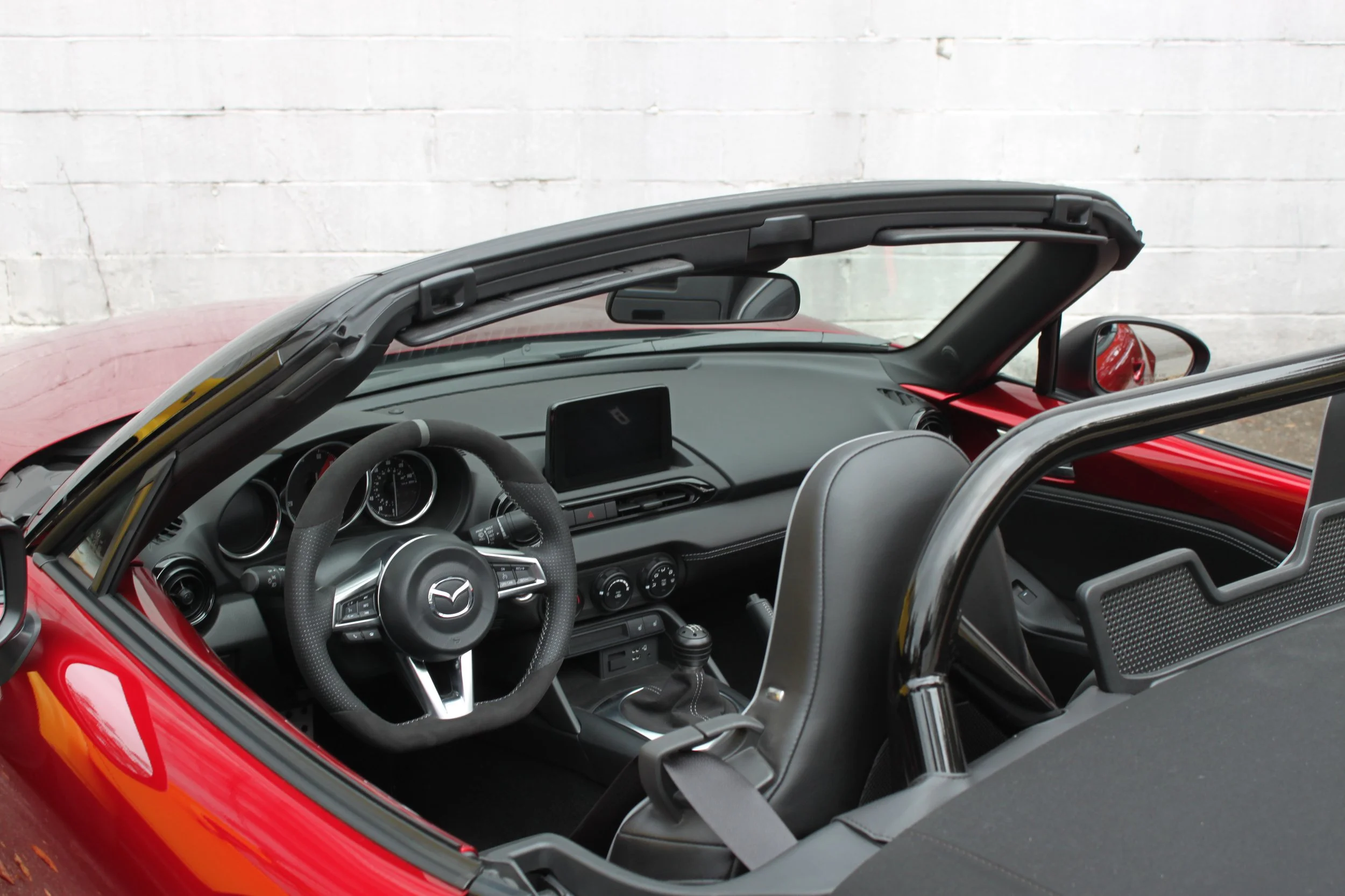 Interior of a red Mazda convertible sports car showing the steering wheel, dashboard, center console with gear shifter, and the driver's seat with a seatbelt, against a white brick wall background.