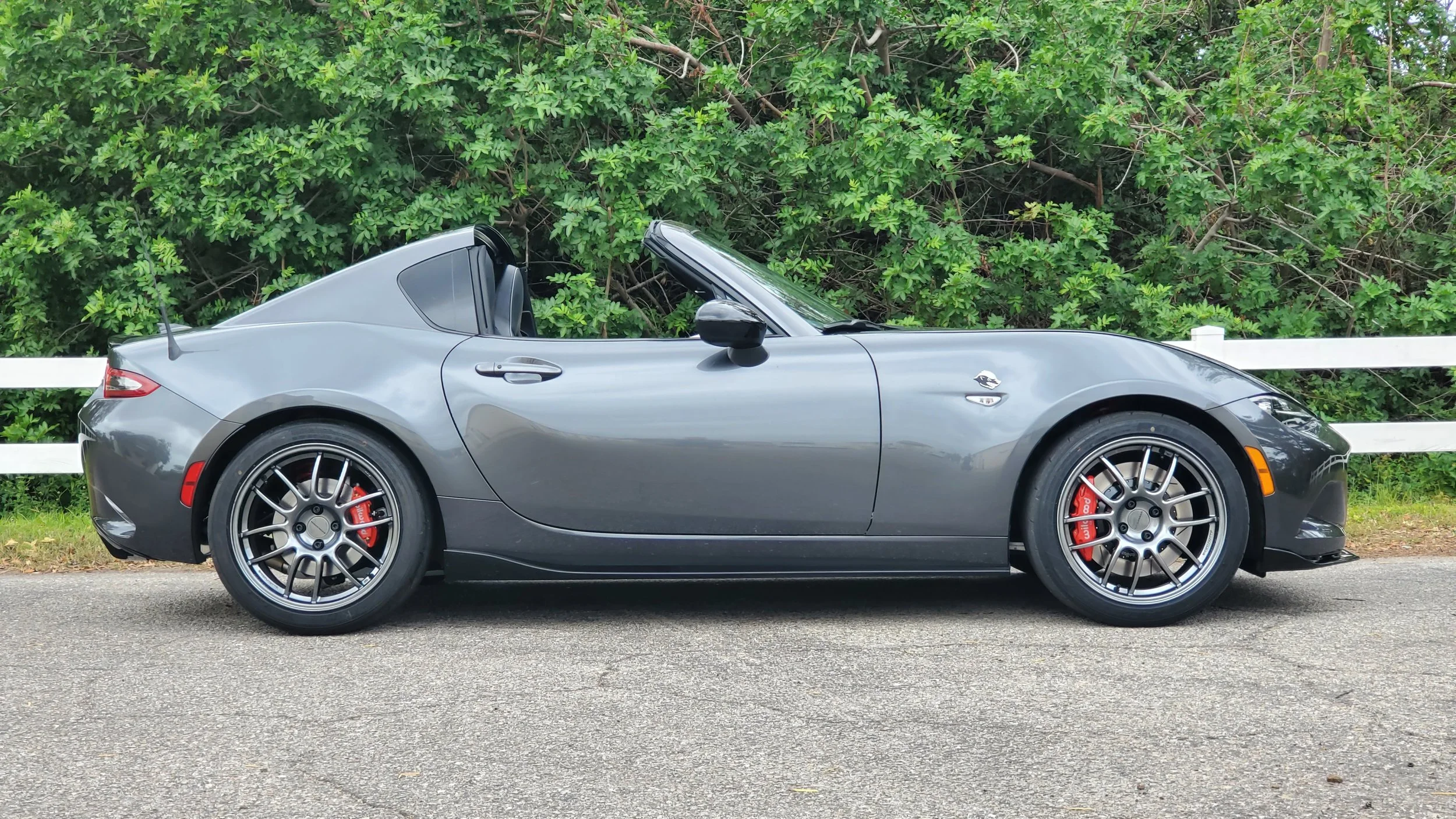 A gray convertible sports car parked on the side of the road with a white fence and lush green bushes in the background.
