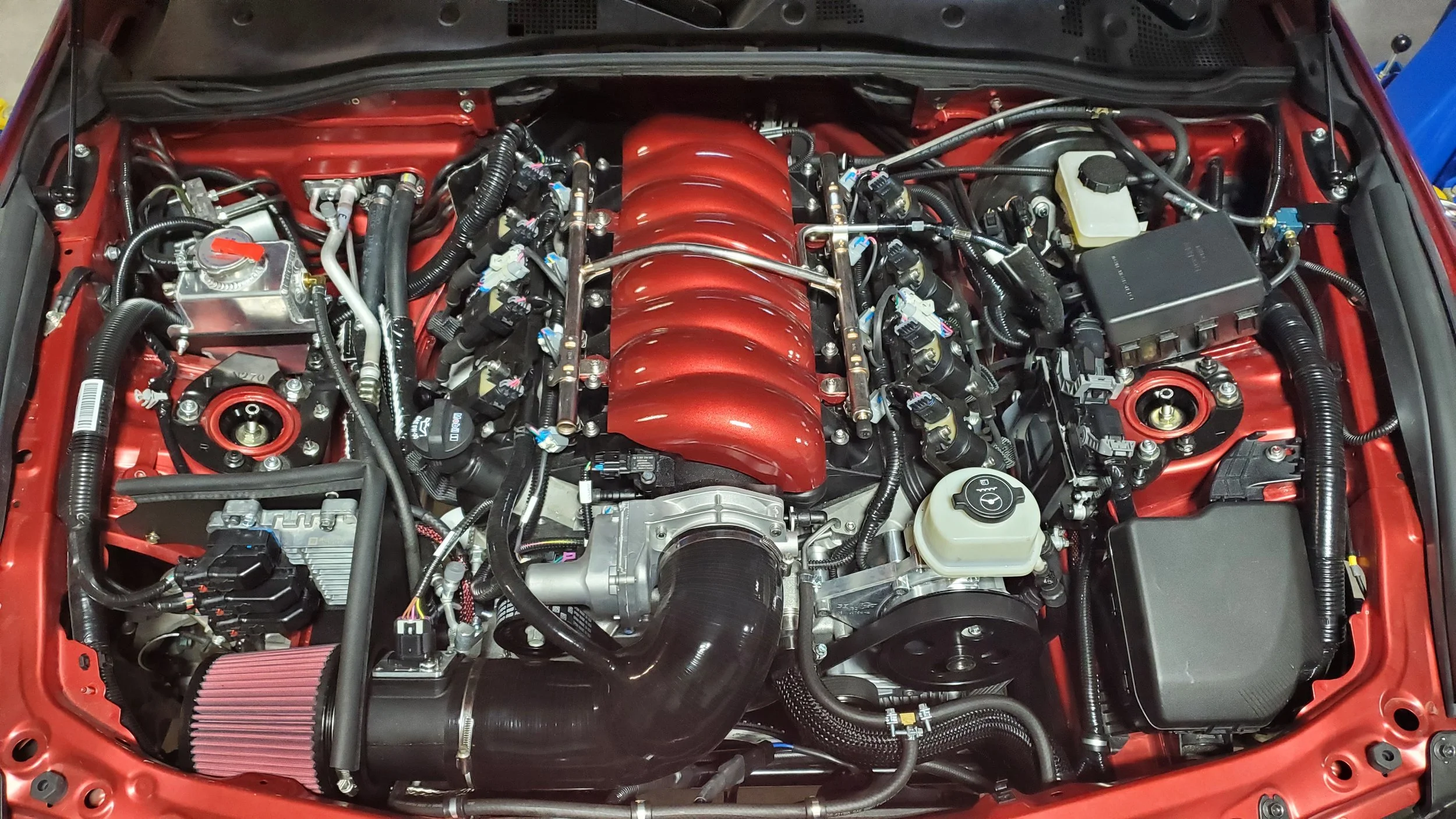 A detailed view of a high-performance car engine, with various components, hoses, and wiring visible, including a red intake manifold and air filter.