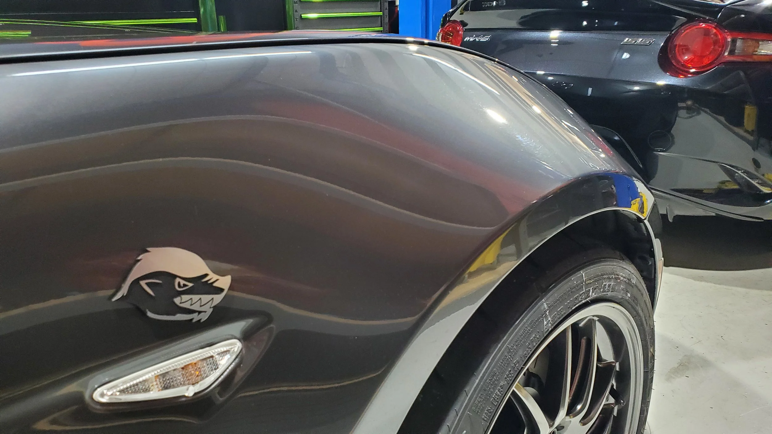 Close-up of a black sports car with a dragon sticker on the front fender, in a garage with other cars in the background.