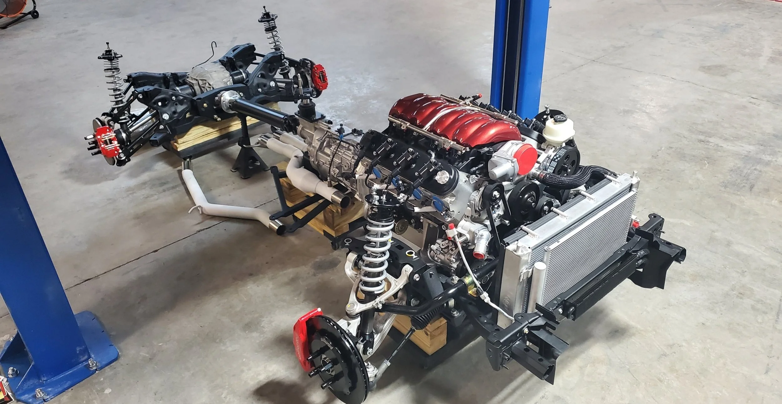 Car chassis and engine assembly on the floor with tools and equipment in the background.