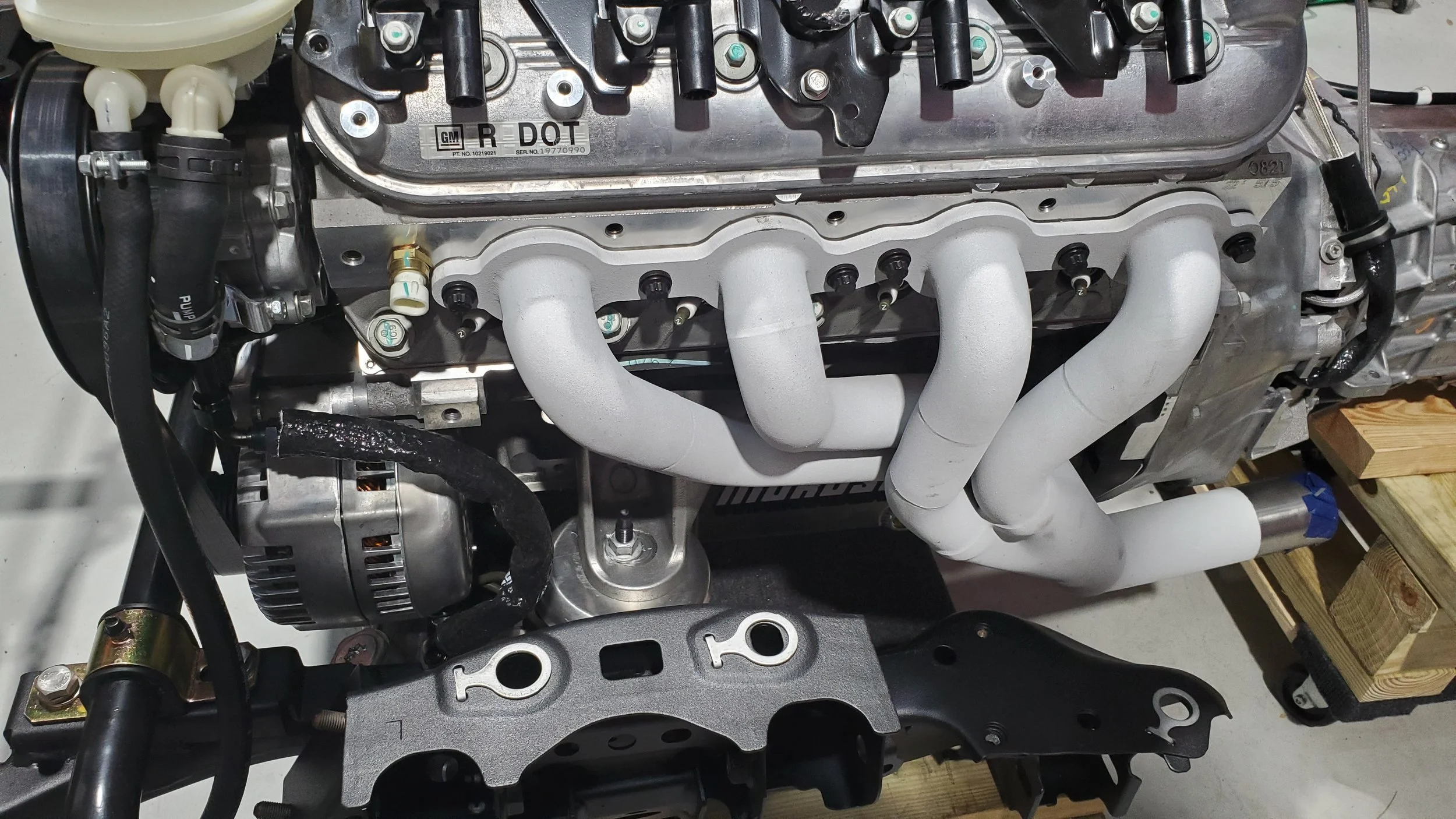 A close-up view of a car engine with white intake manifolds, an alternator, various hoses, and parts, mounted on a black frame, with a wooden platform nearby.