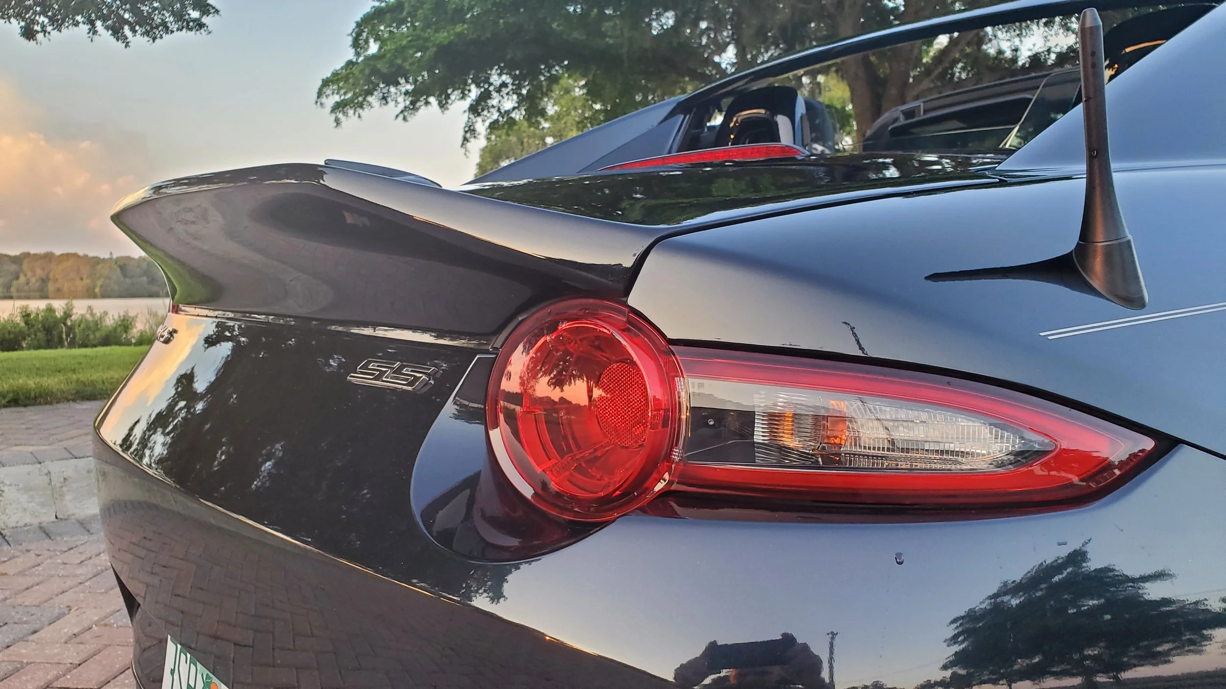 Rear end of a black sports car with a spoiler, taillight, and antenna, parked outdoors near a body of water with trees and a cloudy sky in the background.
