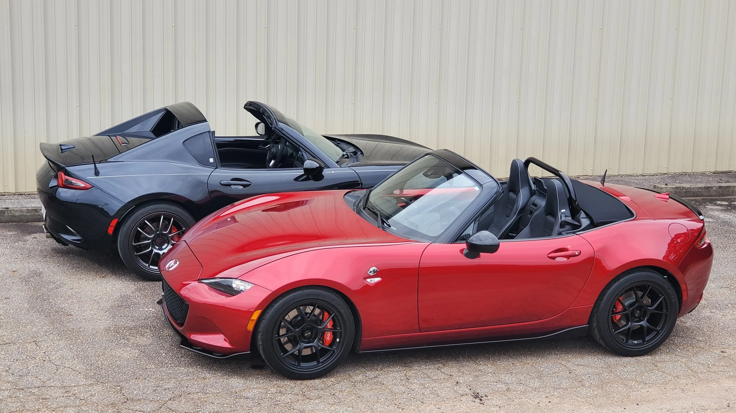 Two Mazda MX-5 Miata convertibles parked side by side on a gravel lot, one red and one black, with a beige metal building in the background.