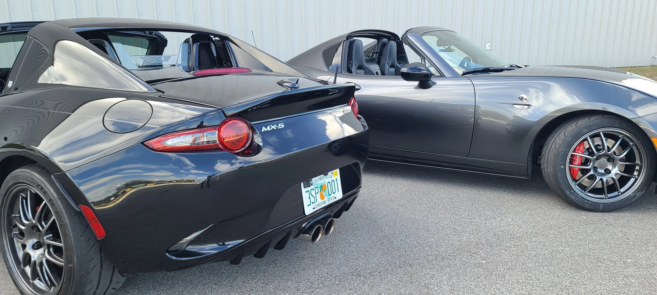 Two sports cars, a black Mazda MX-5 and a gray convertible, parked on an asphalt surface near a beige metal building.