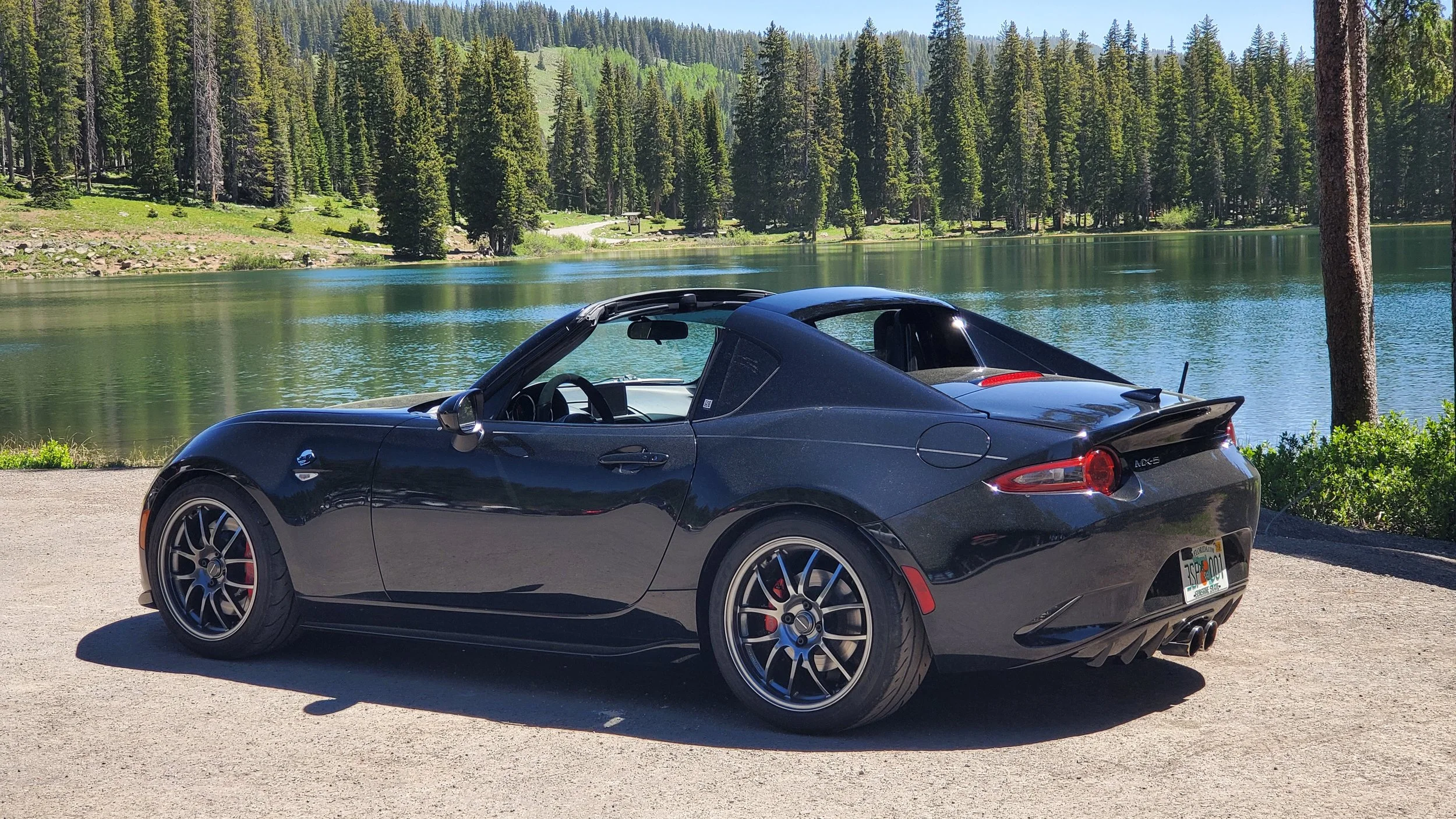 A black sports car parked on a dirt area near a lake, with a backdrop of green pine trees and a forested hillside under a clear blue sky.