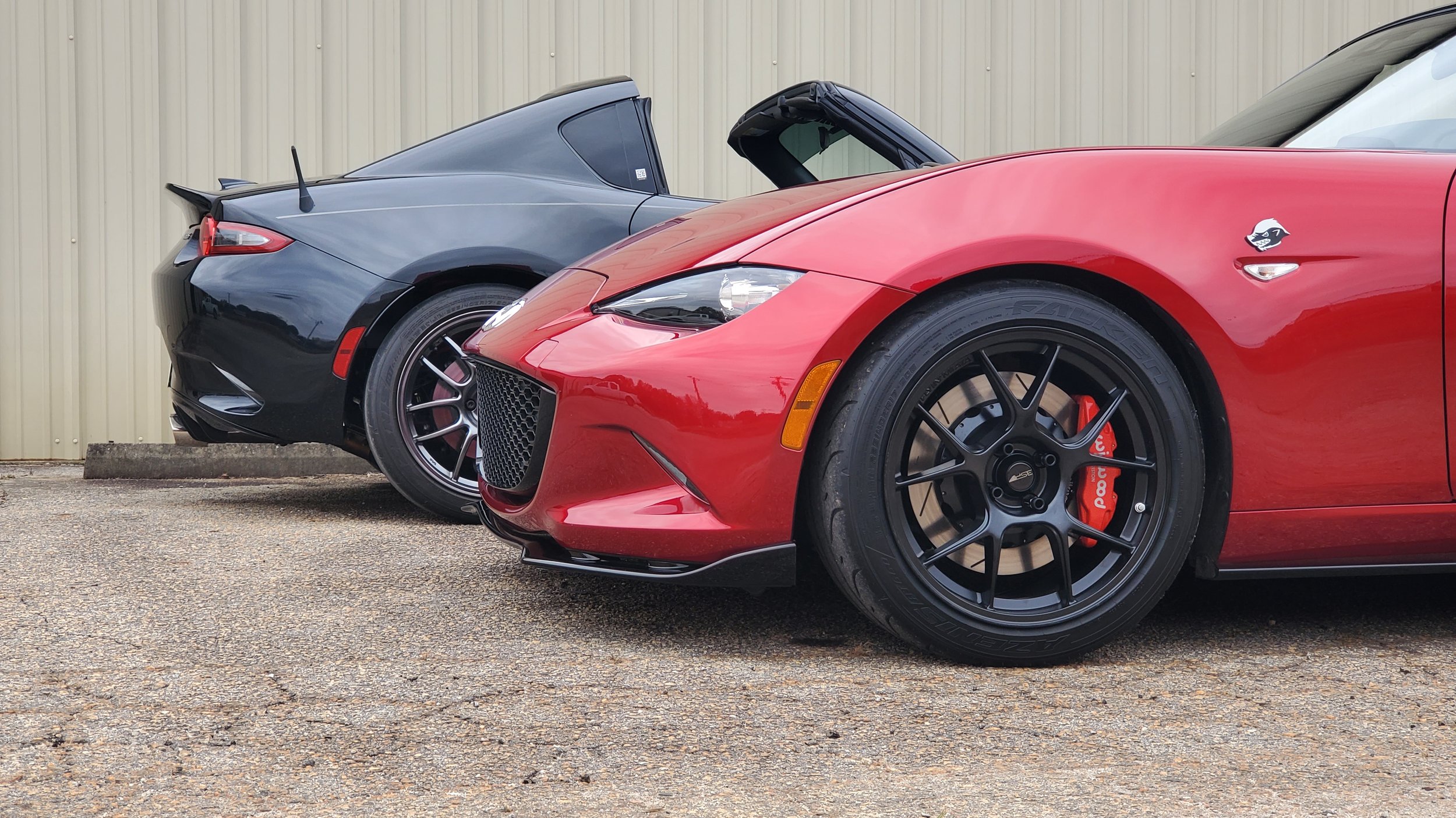 Red sports car with black wheels and red brake calipers, parked next to a black sports car with black wheels, both parked outside in front of a beige metal building.