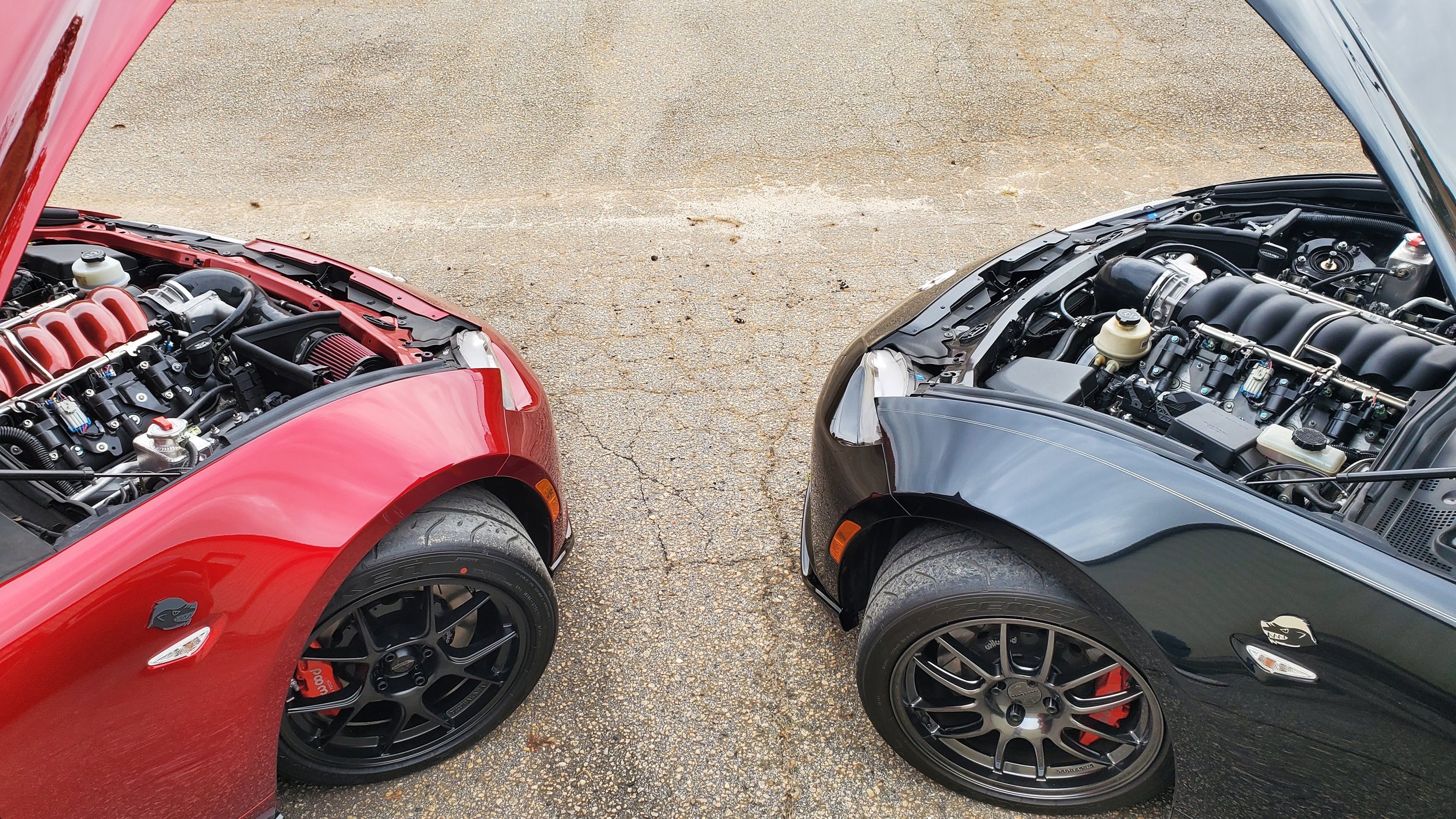 Two sports cars with their hoods open, showing engines, parked on a cracked asphalt surface. The car on the left is red, and the car on the right is black.