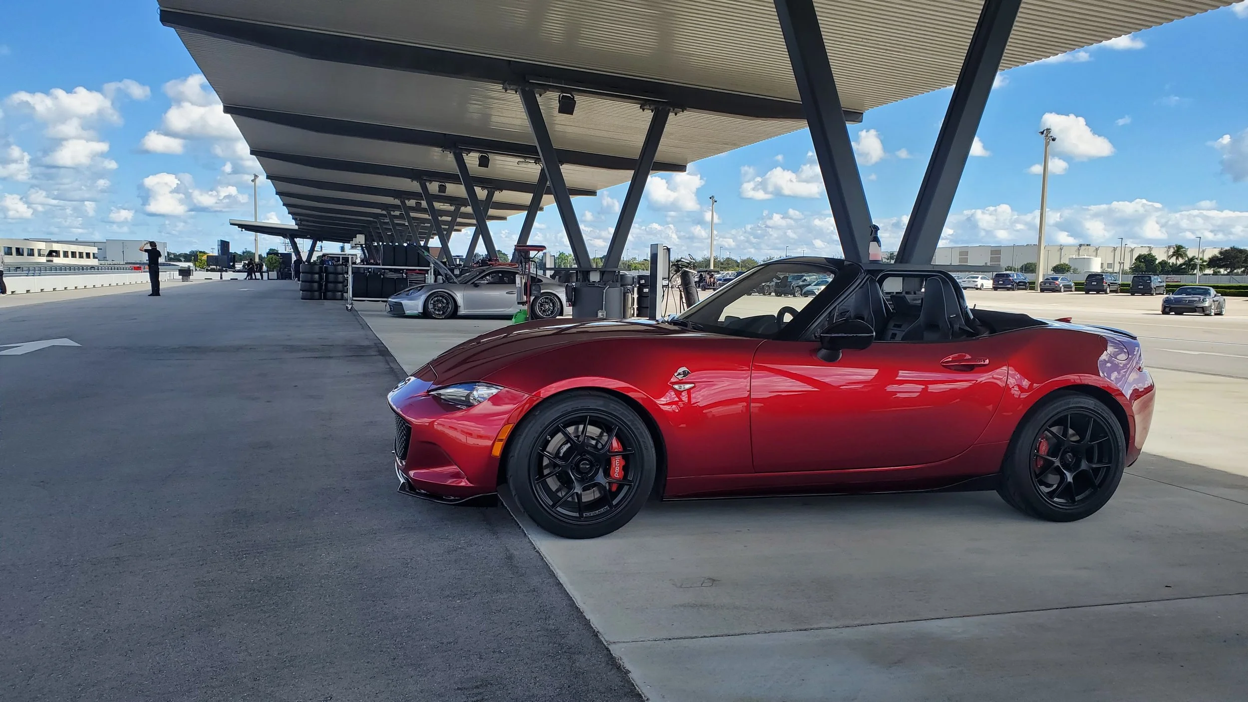 A red sports car parked at a race track or pit area with other cars and a person in the background, under a large, modern canopy with a clear blue sky and some clouds.