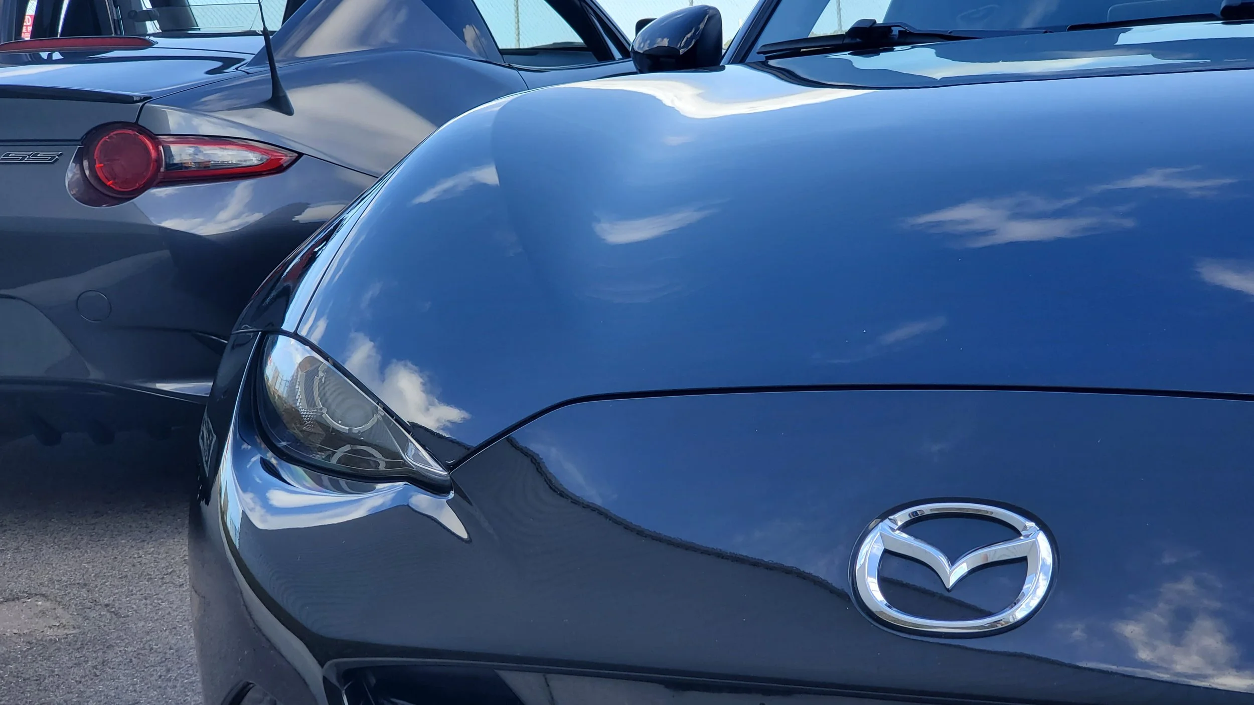 Close-up view of the front of a black Mazda car, showing the headlight, hood, Mazda emblem, and part of the windshield with a reflection of clouds. Behind it, there is a gray sports car with an orange tail light.